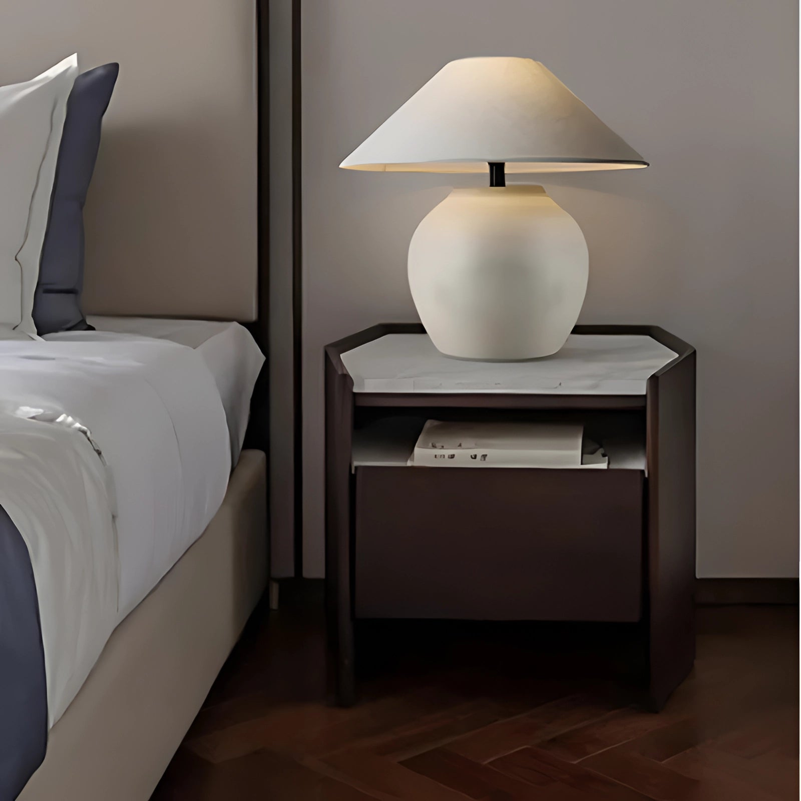 Modern dark wood bedside table with a minimalist design featuring a rectangular shape, a built-in shelf, and a lamp placed on top, set against a grey and white interior with a wooden floor.