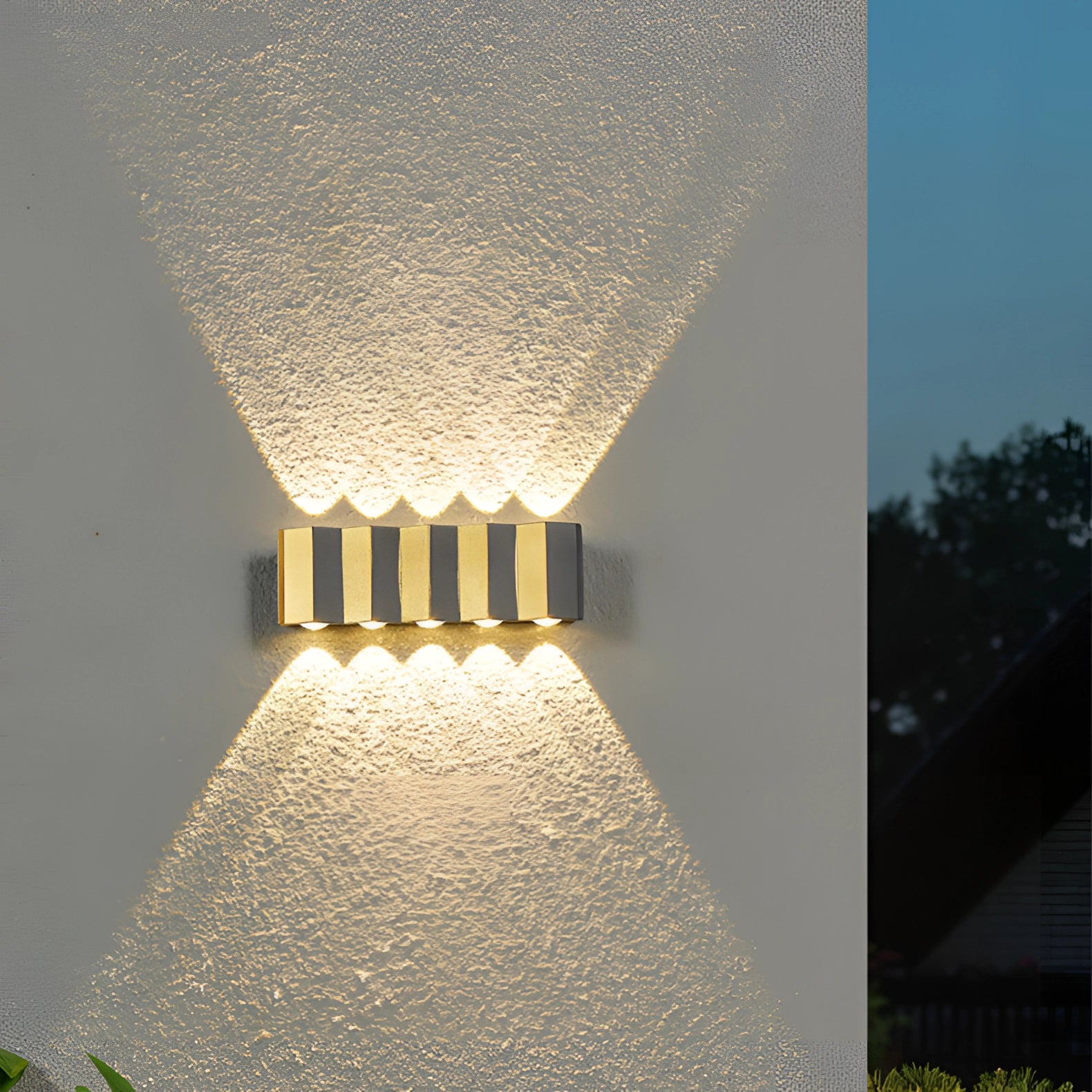 Modern geometric LED wall lamp mounted on an outdoor wall, featuring a sleek rectangular design. The fixture is set against a natural background with plants and trees visible, highlighting its use in an outdoor setting. The lamp emits a soft glow, creating artistic tints and shades on the wall.