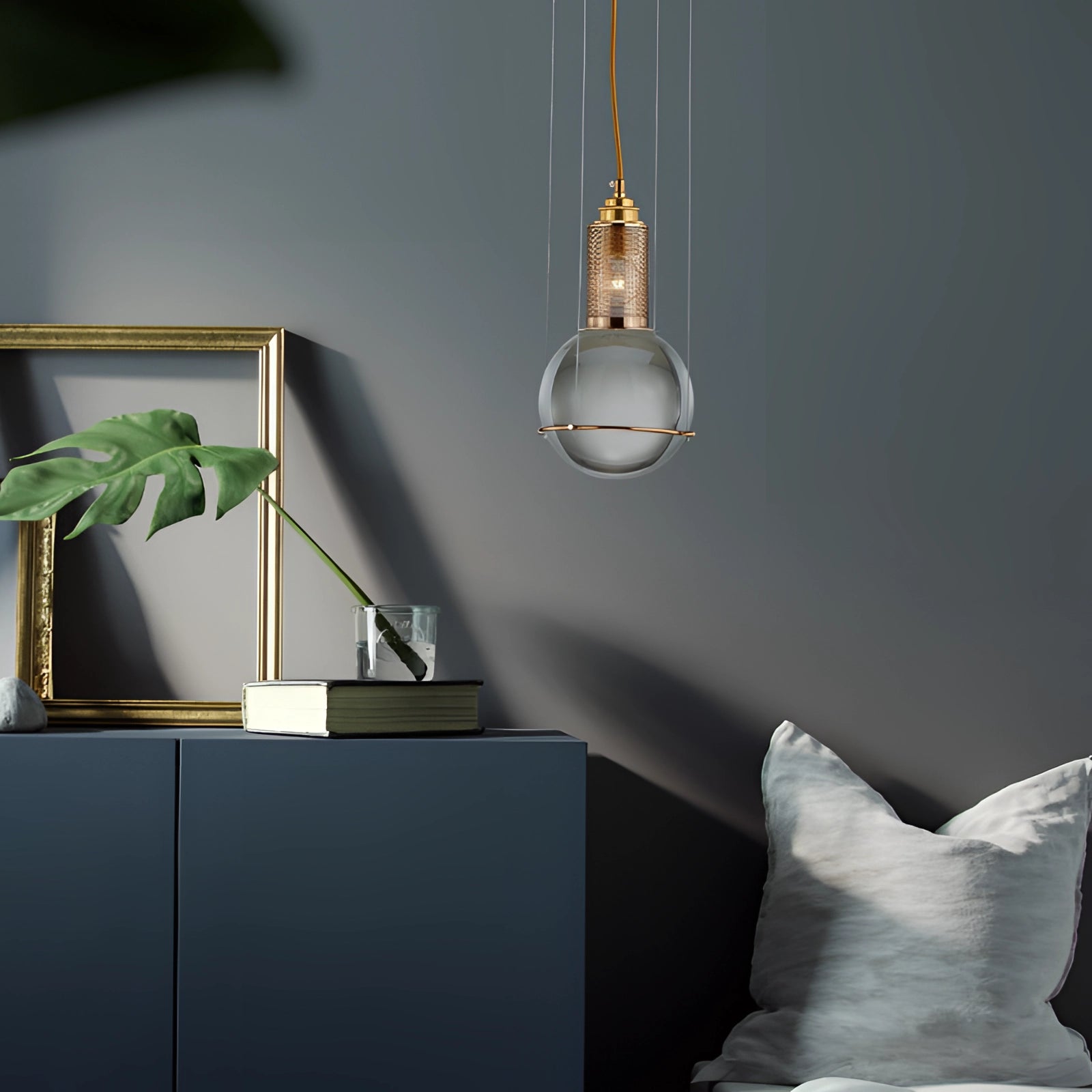 Modern glass globe pendant light with a gold finish hanging from a ceiling in a minimalist interior design setting, surrounded by wooden shelving and houseplants.