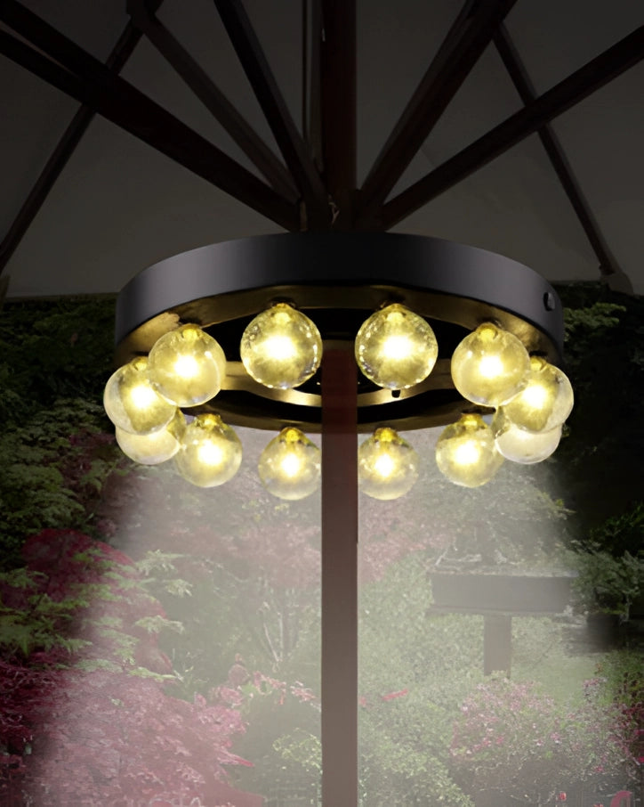 Modern industrial round chandelier with exposed filament bulbs hanging in a decorated room with plants and a tree in the background.