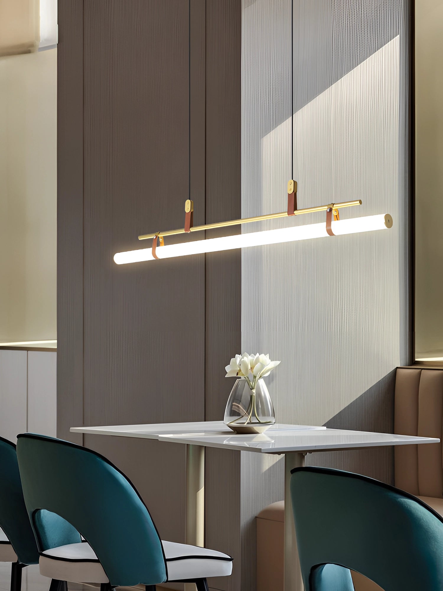 Modern minimalist kitchen and dining room featuring a sleek LED tube pendant light hanging above a contemporary wooden table surrounded by chairs, with shelving and a potted plant enhancing the stylish interior design.