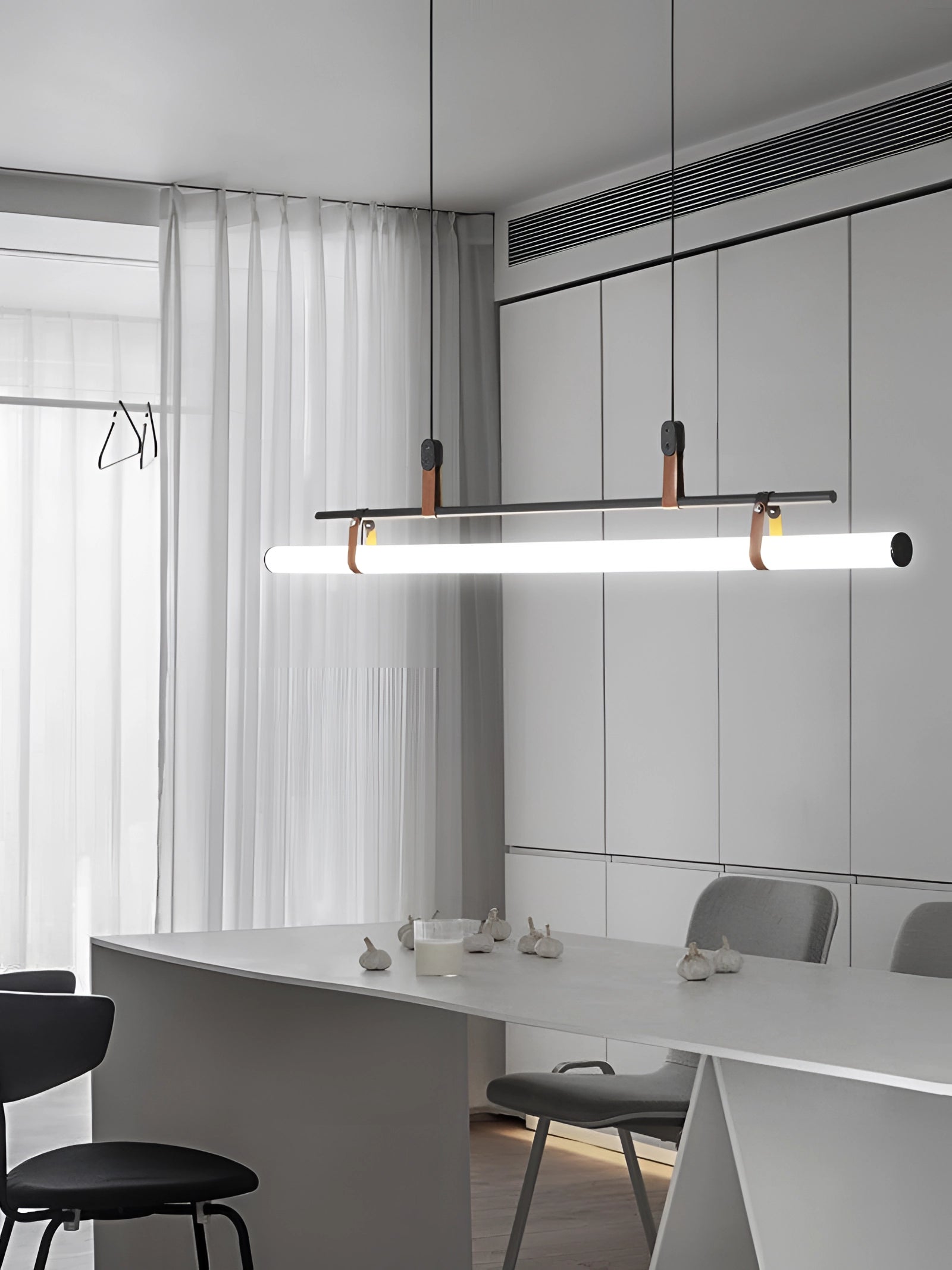 Modern LED tube pendant light hanging above a minimalist kitchen and dining room setting featuring sleek wooden furniture, including a table and chairs, with soft curtains and neutral-colored walls in the background.