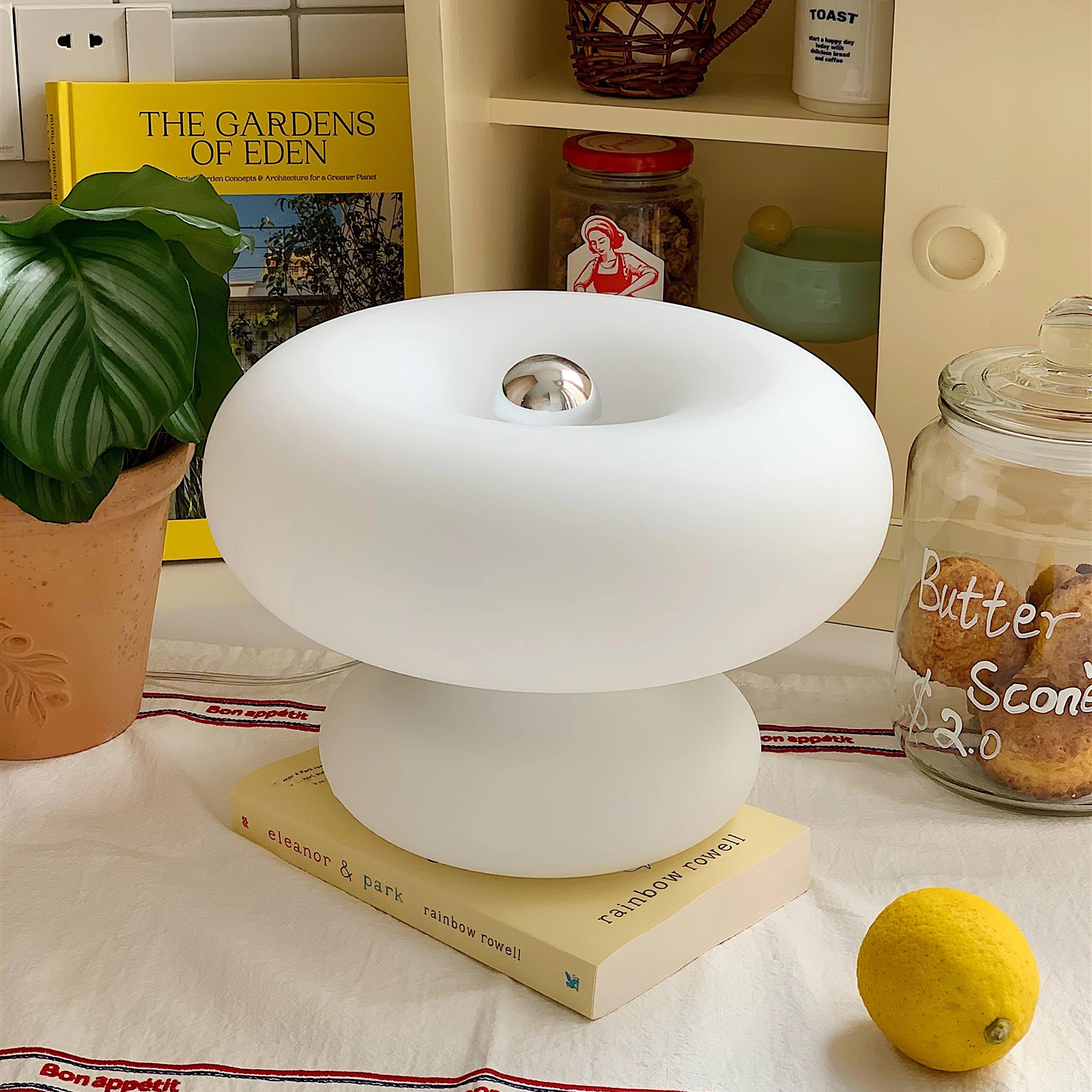 A sleek, modern mushroom-shaped table lamp with a minimalist design, placed on a shelf beside a small potted plant and decorative items.