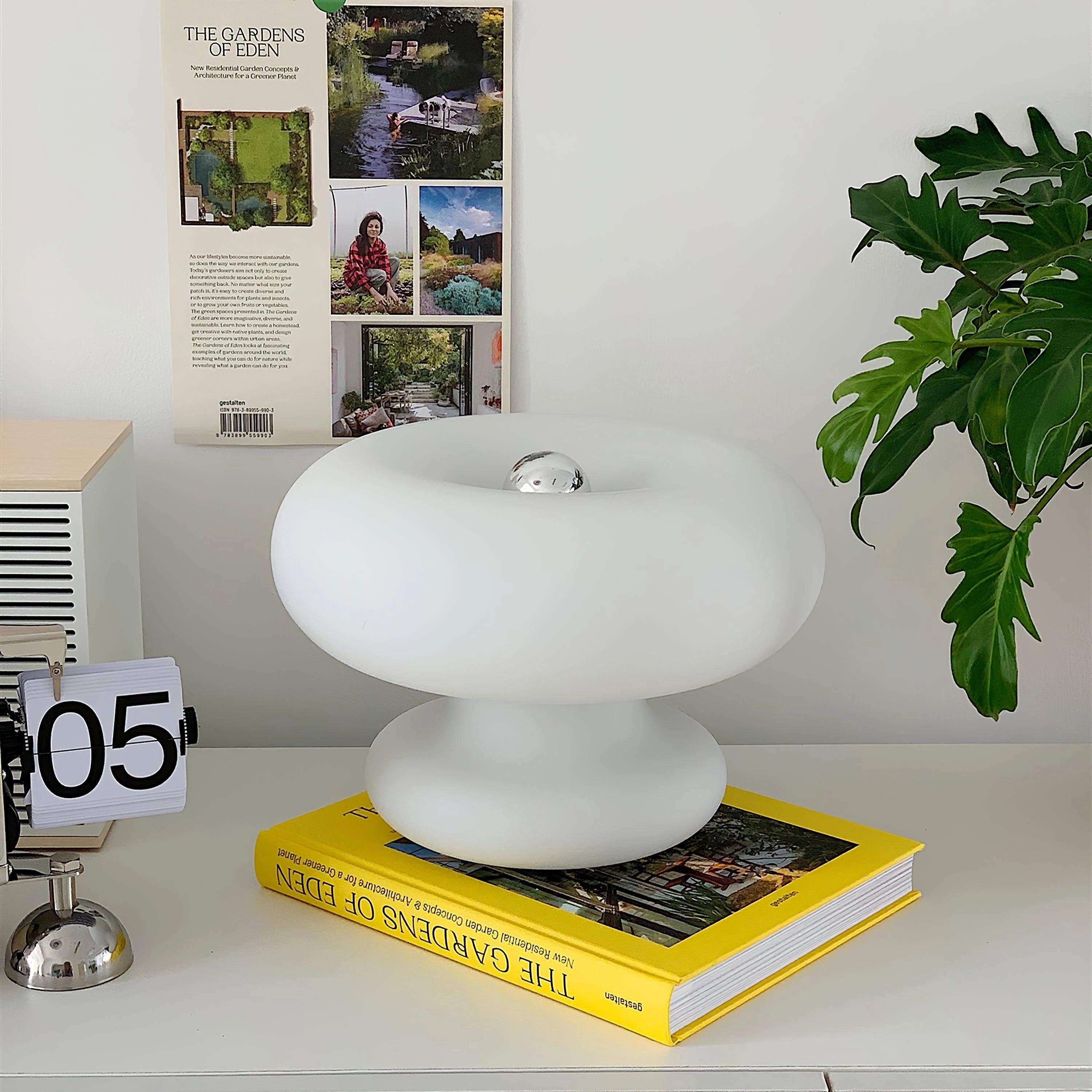 Modern mushroom table lamp with a minimalist design, placed on a sleek table in a stylish living room setting, accompanied by a plant and shelving décor.