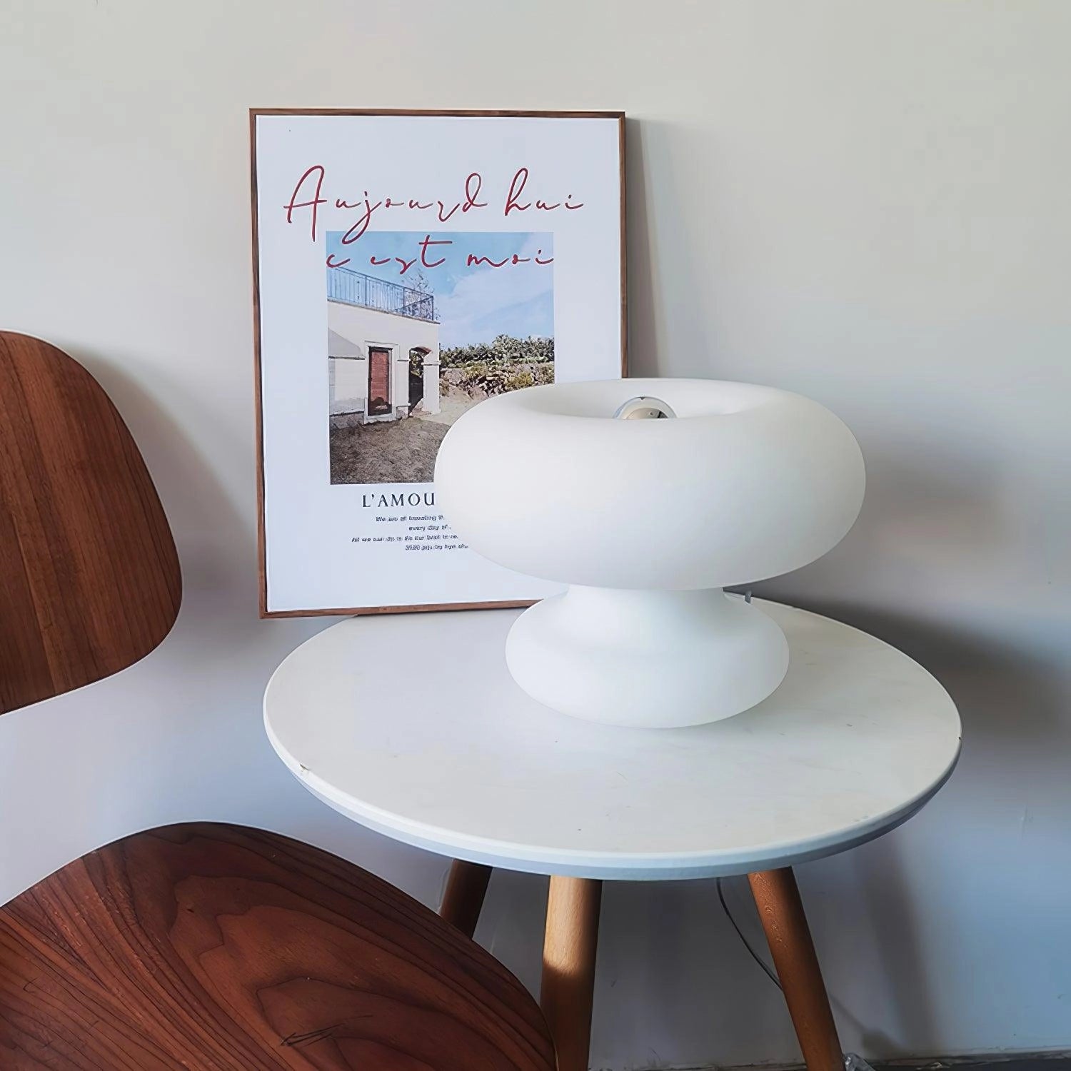 A modern mushroom-shaped table lamp with a minimalist design, placed on a wooden bedside table with white tableware and a chair nearby.