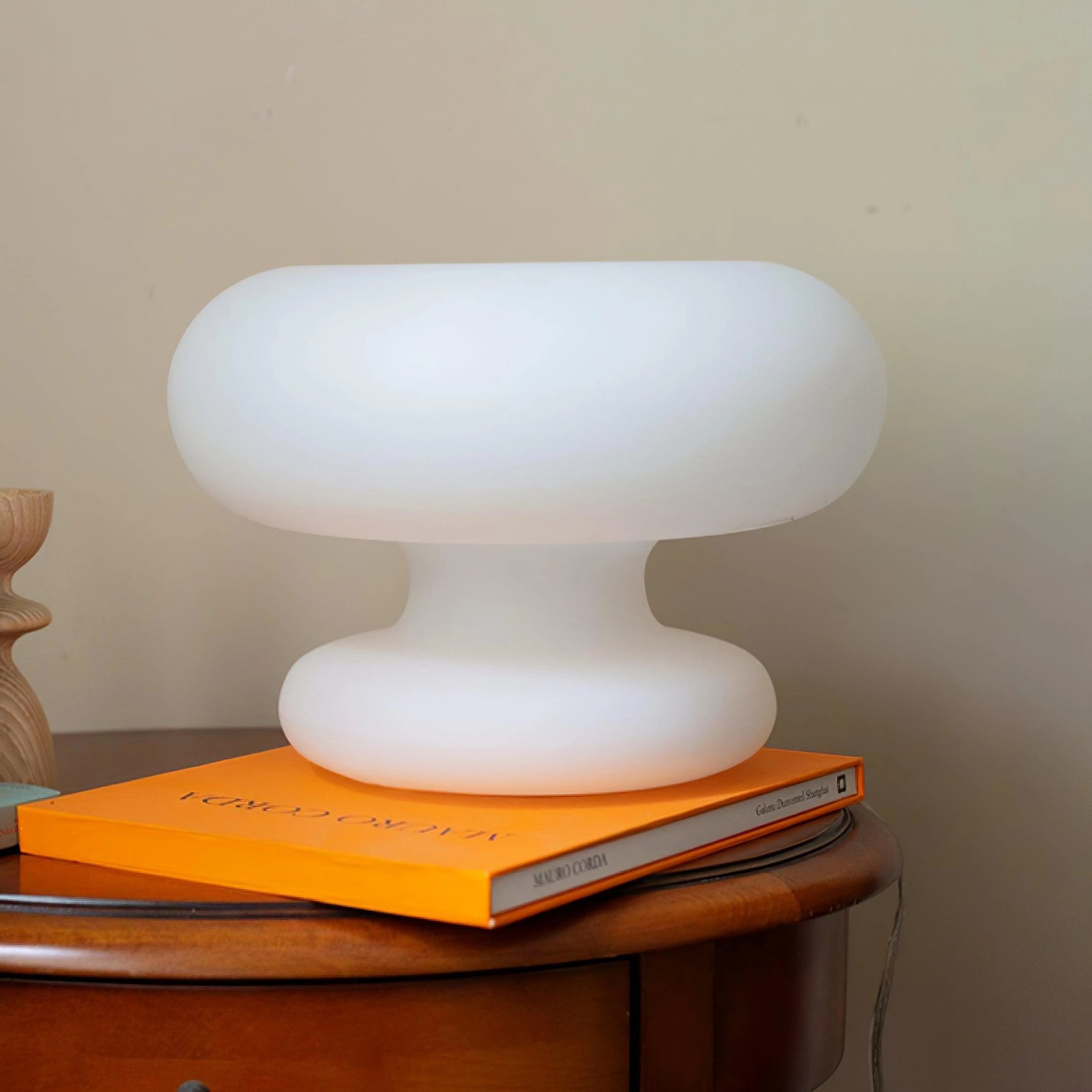 A minimalist designer bedside light shaped like a mushroom, placed on a wooden table with a shelf in the background.