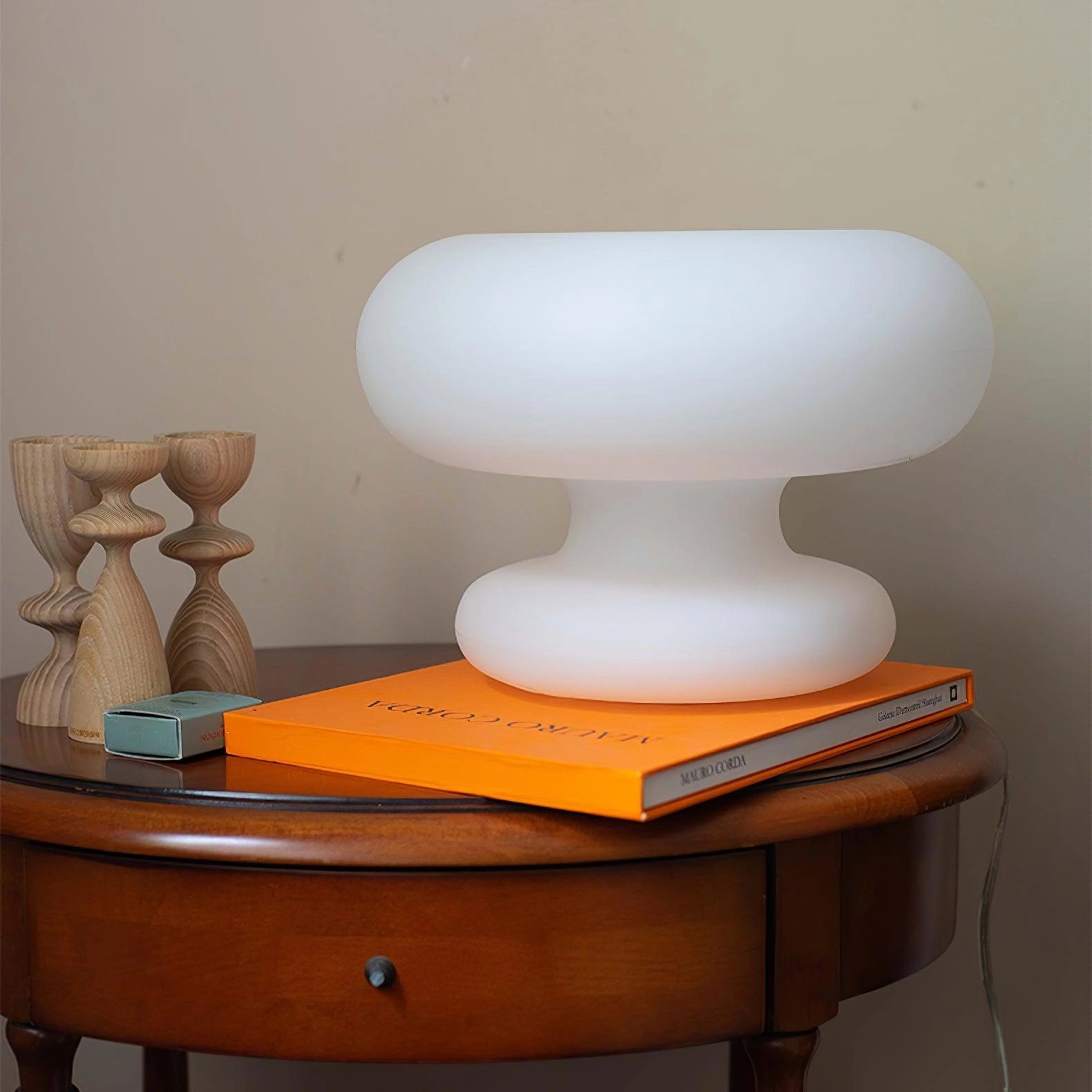 Modern mushroom-shaped table lamp with minimalist design, placed on a wooden desk near a chair and shelving unit. The lamp features a sleek, varnished finish, ideal for a bedside or modern living space.