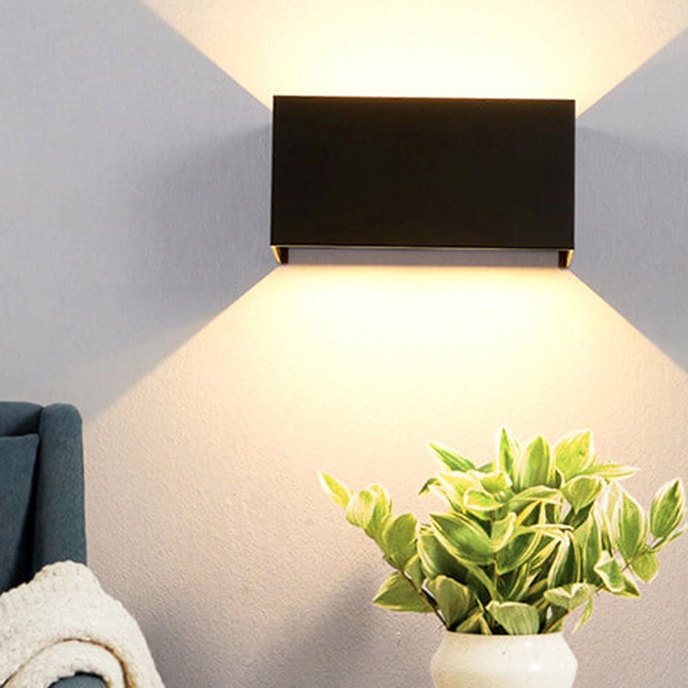 Modern rectangular black LED outdoor wall light mounted on a white wall, surrounded by potted houseplants and a wooden table.