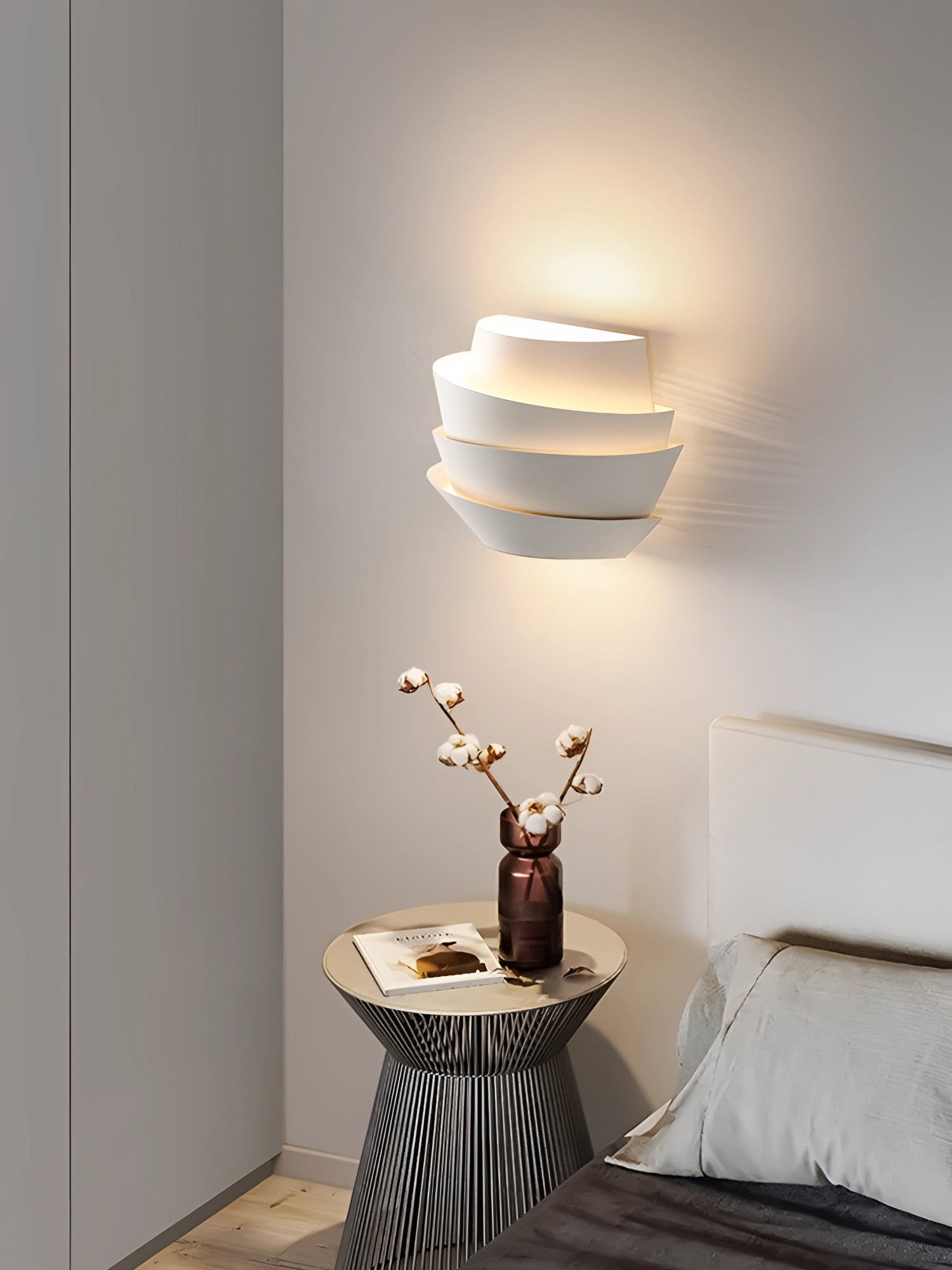 Modern sculptural LED pendant light with a minimalist spiral design, suspended above a wooden table that features a small plant, complementing the clean, white and grey interior with artistic wall elements in the background.