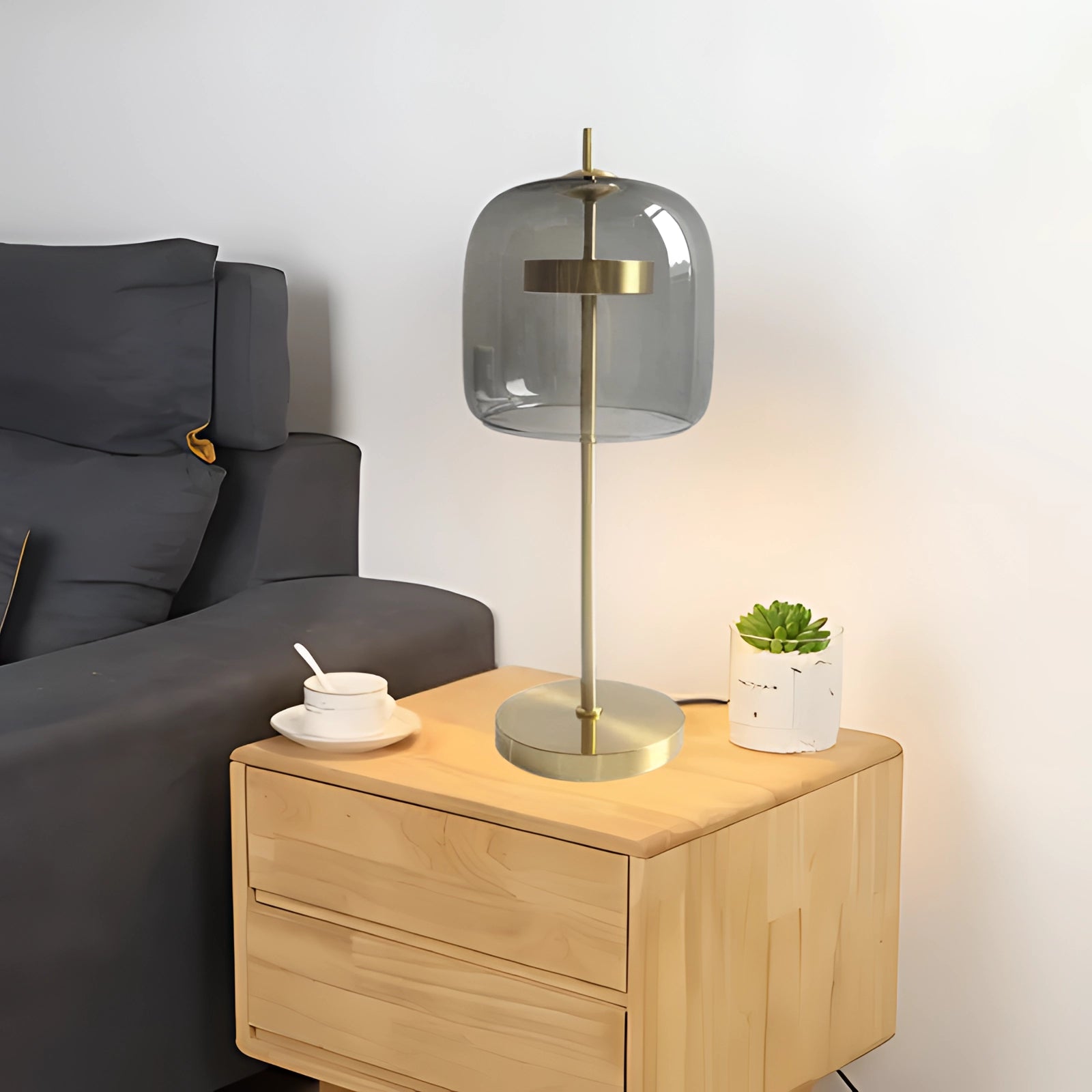 Modern smoked glass lamp with brass accent and gold base on a wooden floor, surrounded by furniture and interior decor, including a chair and a plant, with shelving in the background.