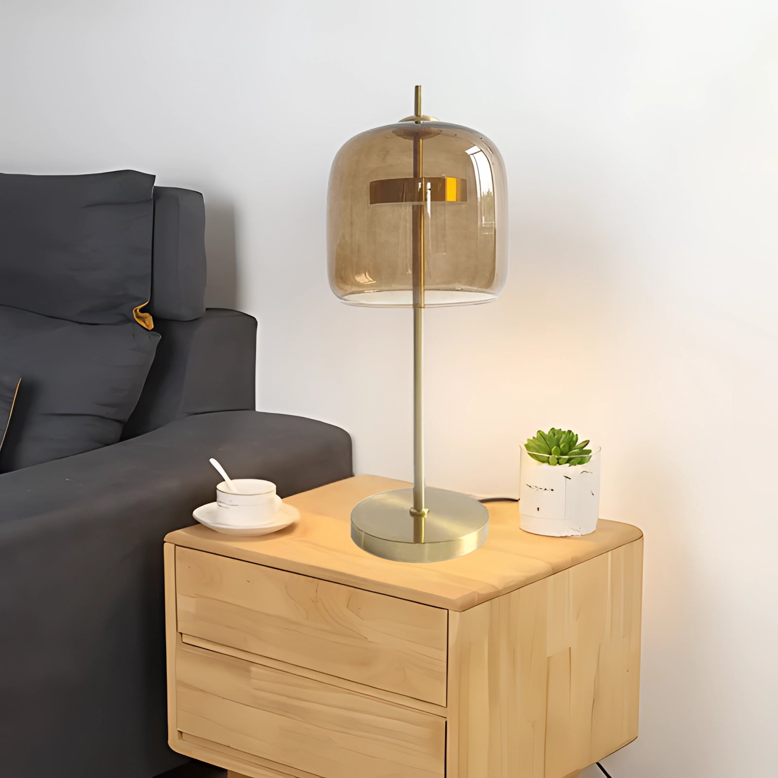 A modern smoked glass lamp with a brass accent and a gold base, featuring an amber shade and emitting a cold white light, placed on a wooden shelf next to a potted plant in a stylish interior setting.