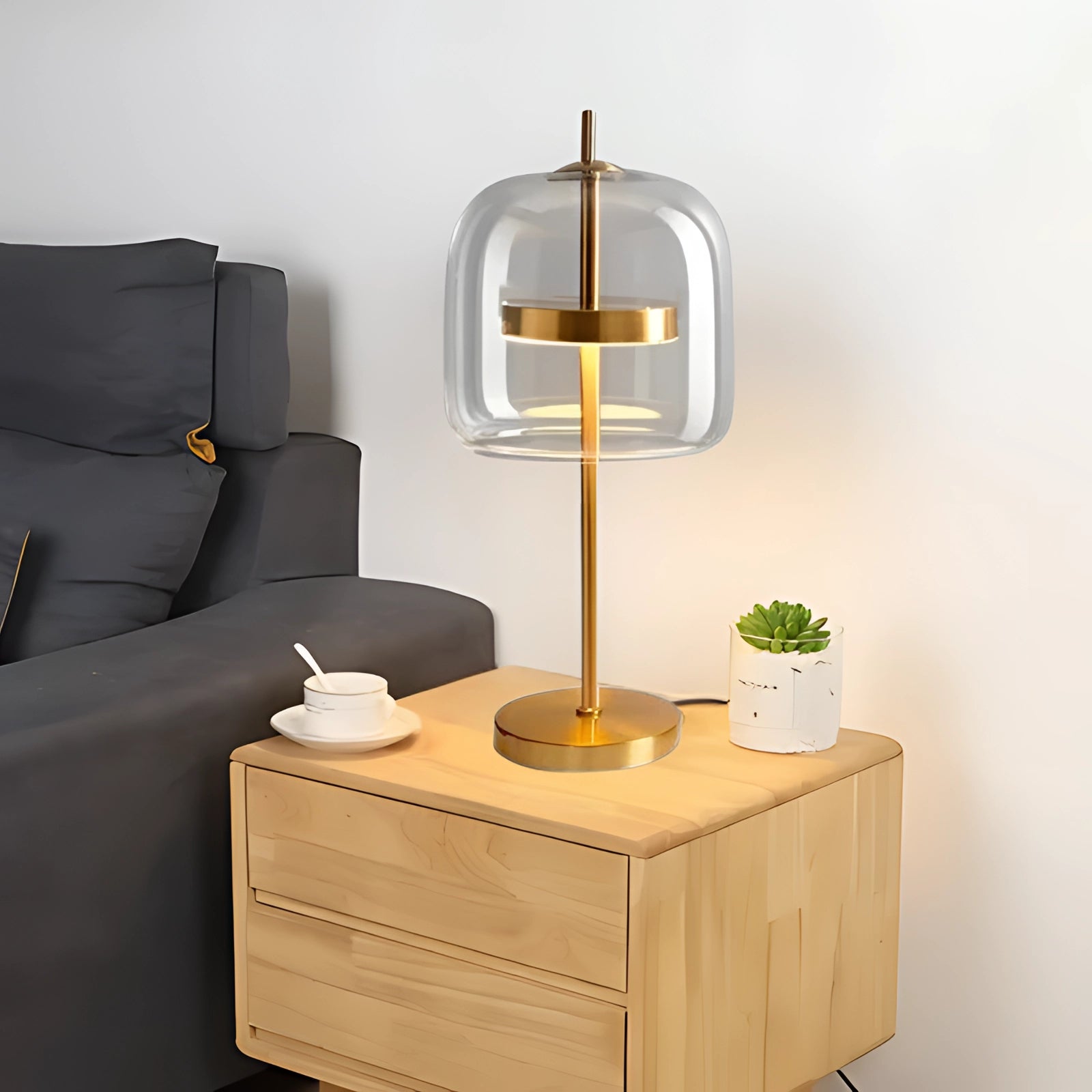 Modern smoked glass lamp with brass accent featuring a transparent shade and a gold base, placed on a wooden shelf in a contemporary interior setting with cold white lighting.