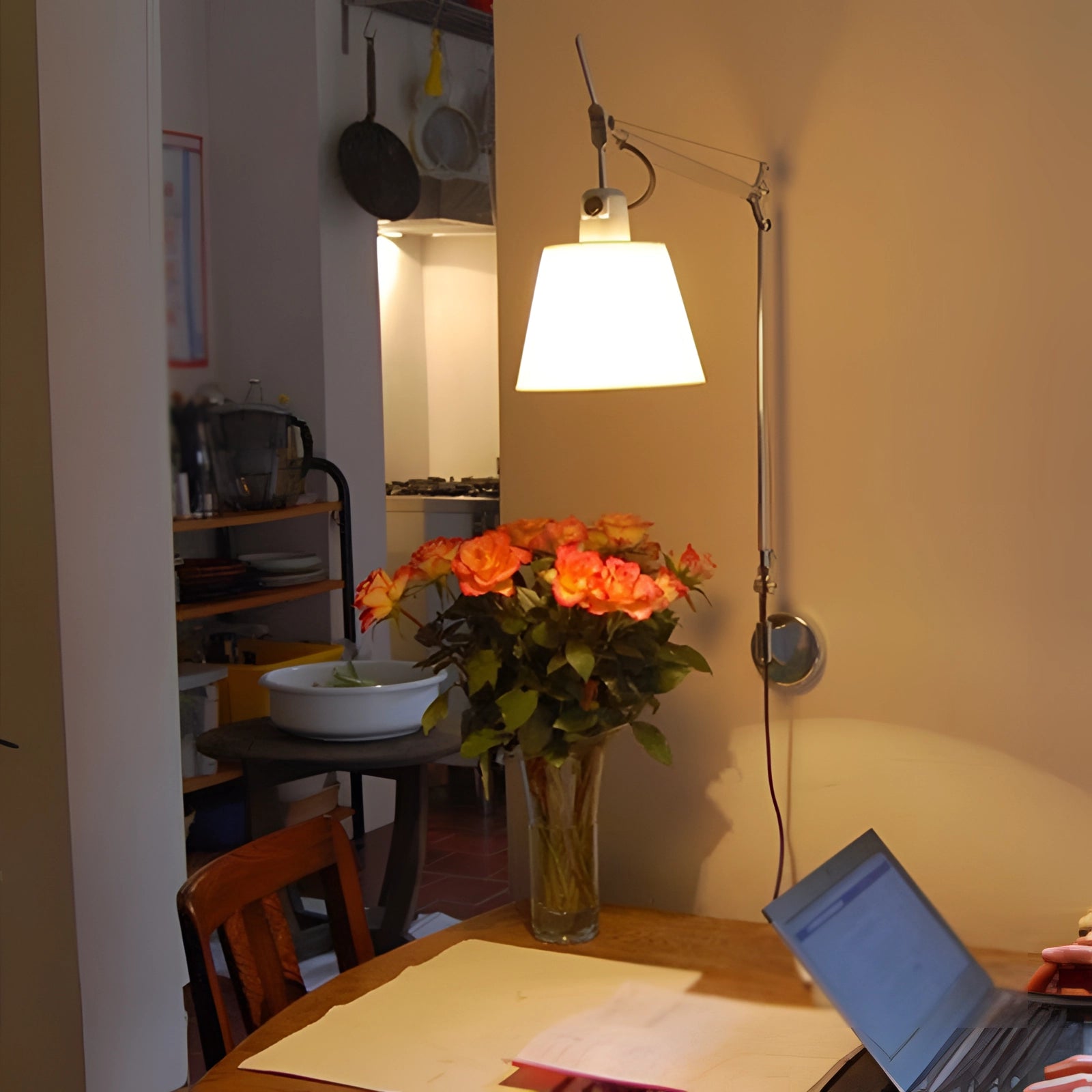 Modern silver swing arm wall-mounted lamp with shade illuminating a workspace area featuring a table with a laptop, a small houseplant, and a decorative flower, providing adjustable lighting for an elegant and functional setting.