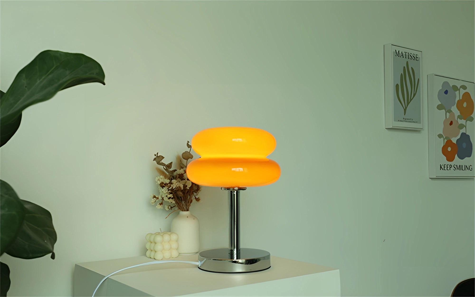 Modern tiered glass table lamp with a minimalist design placed on a wooden surface in a living room setting, surrounded by a lush green houseplant in a flowerpot. The lamp features a peach-toned lampshade, complementing the interior design, with art visible on the walls and sleek flooring in the background.