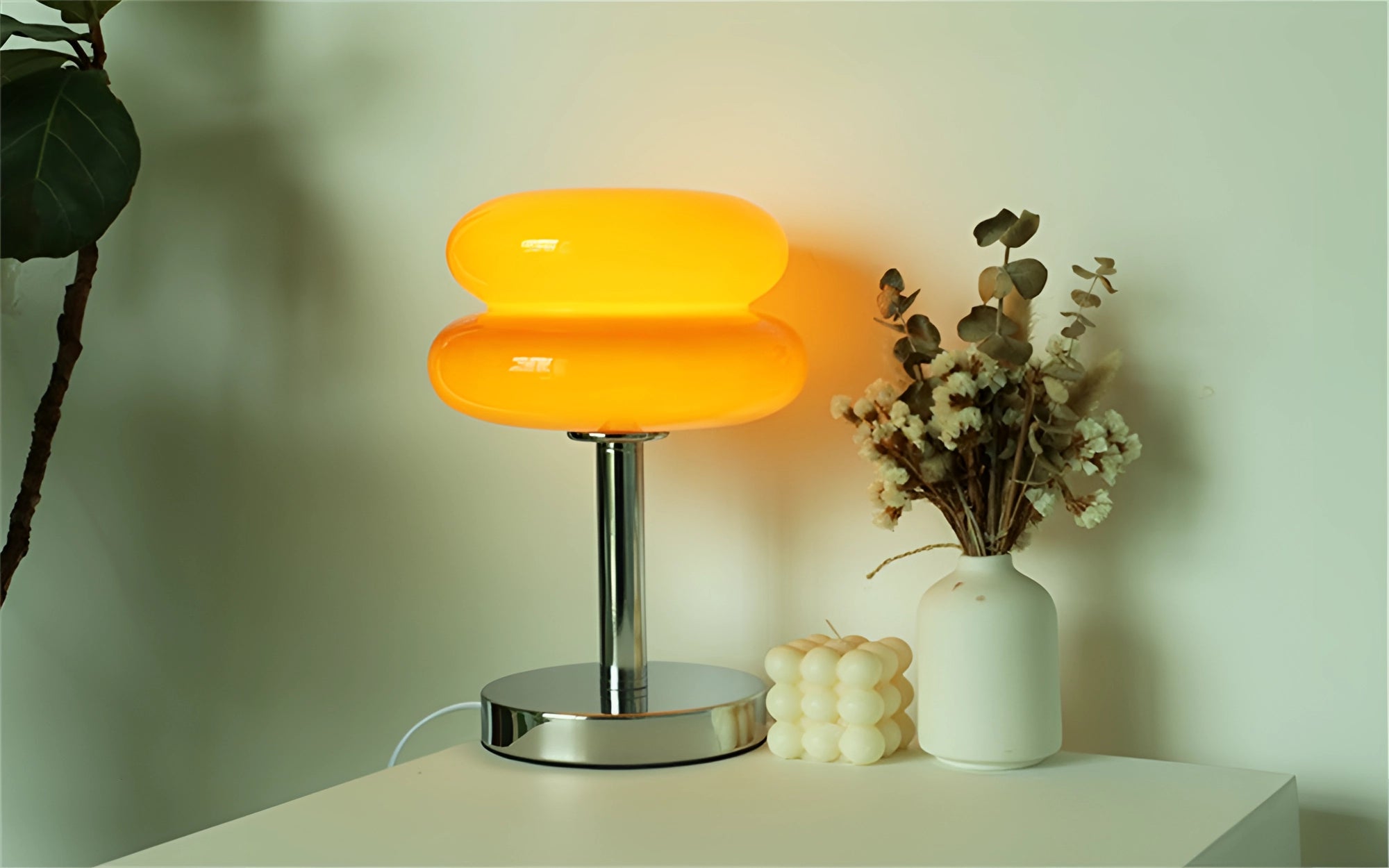 Orange-colored modern tiered glass table lamp with thick bottom, placed on a wooden table beside a green houseplant in a vase, designed for minimalist bedside or living room interior.