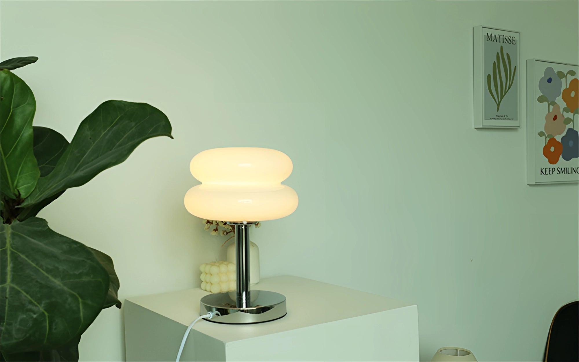 Modern glass table lamp with a tiered design placed on a wooden shelf in a minimalist living room setting, accompanied by a green plant and neutral-toned decor elements.