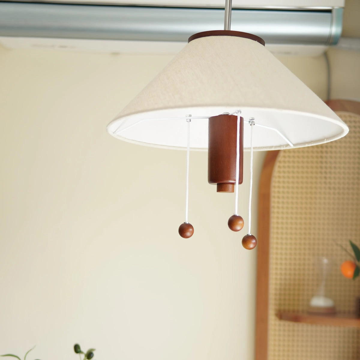 Modern triangle pendant lamp with wooden accents hanging from the ceiling, illuminating a room with a table, plant, and window in the background.