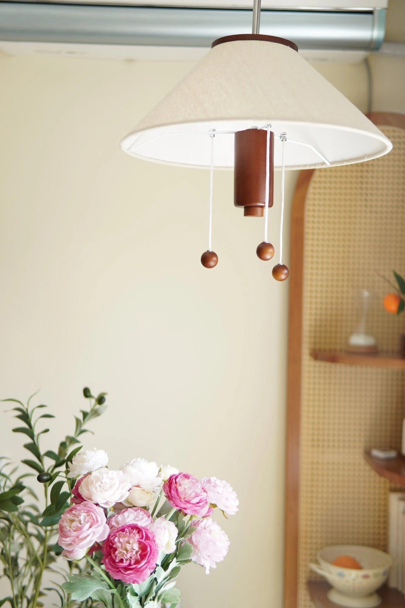 Modern triangle pendant lamp with wooden accents hanging in a stylish interior, surrounded by white and pink flowers and wooden shelving.