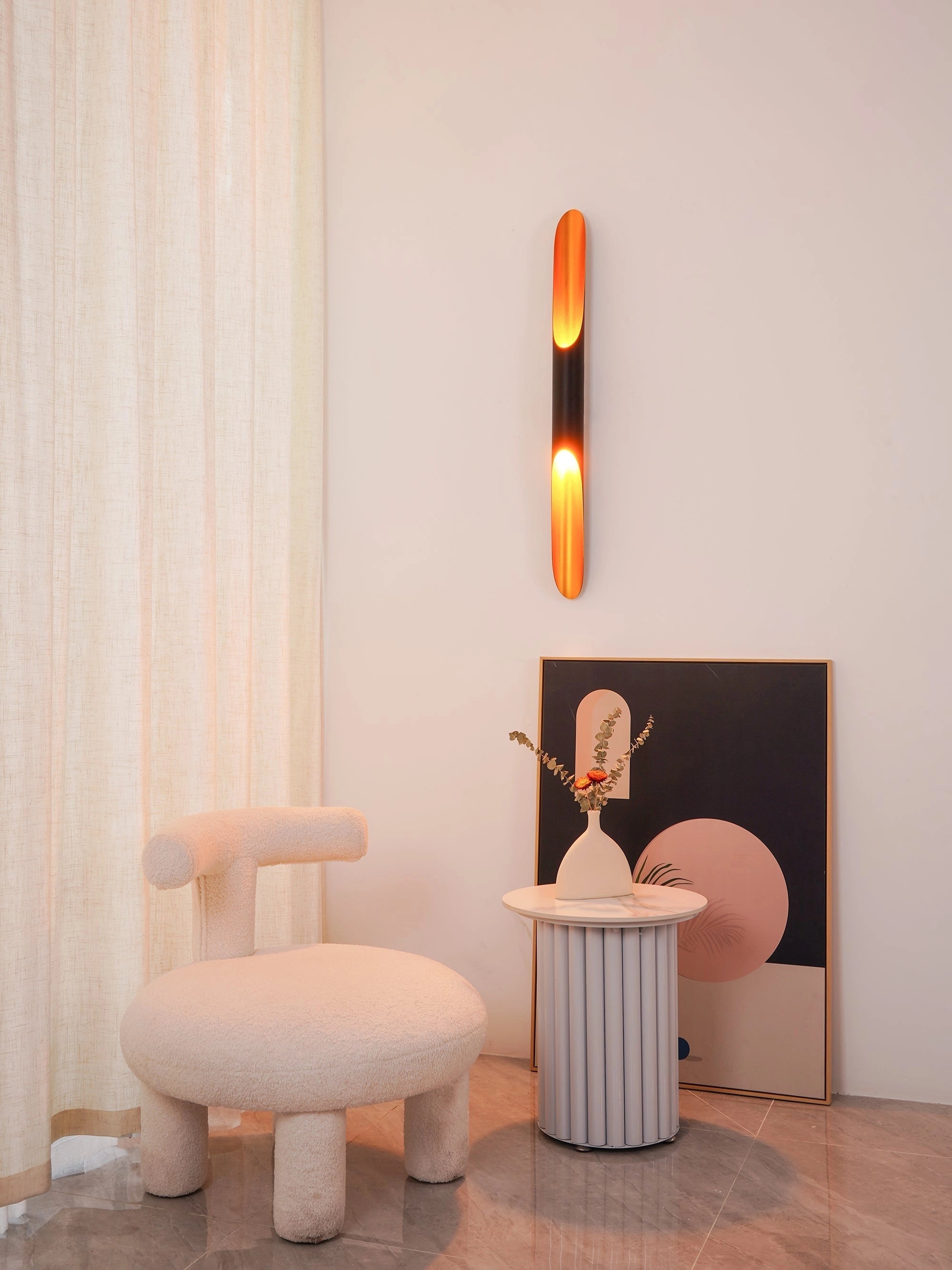 Modern vertical LED wall sconce mounted on a warm-toned wall, illuminating a minimalistic room featuring wooden furniture and an orange stool. The room showcases sleek interior design elements including a wooden floor and a simple ottoman.