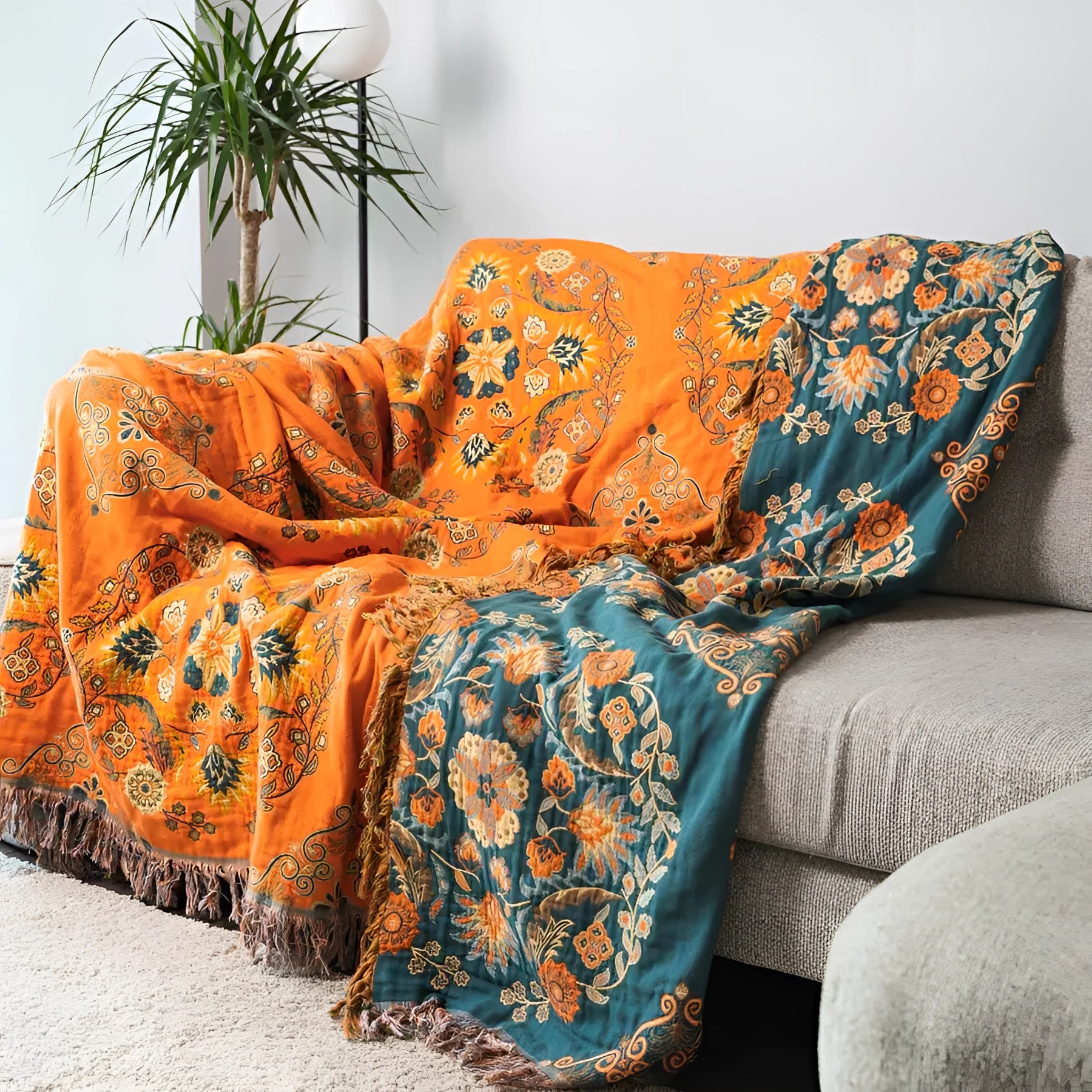 Orange paisley bohemian tassel throw blanket draped over a studio couch, adorned with pillows, next to a houseplant in a flowerpot, emphasizing comfort and luxury cotton decor.