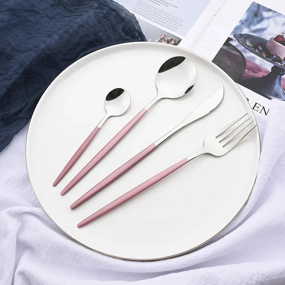 Pink stainless steel flatware set displayed on a table, featuring a modern and elegant design. The set includes forks and other cutlery pieces, complemented by dishware and textile elements, creating an inviting dining setting.