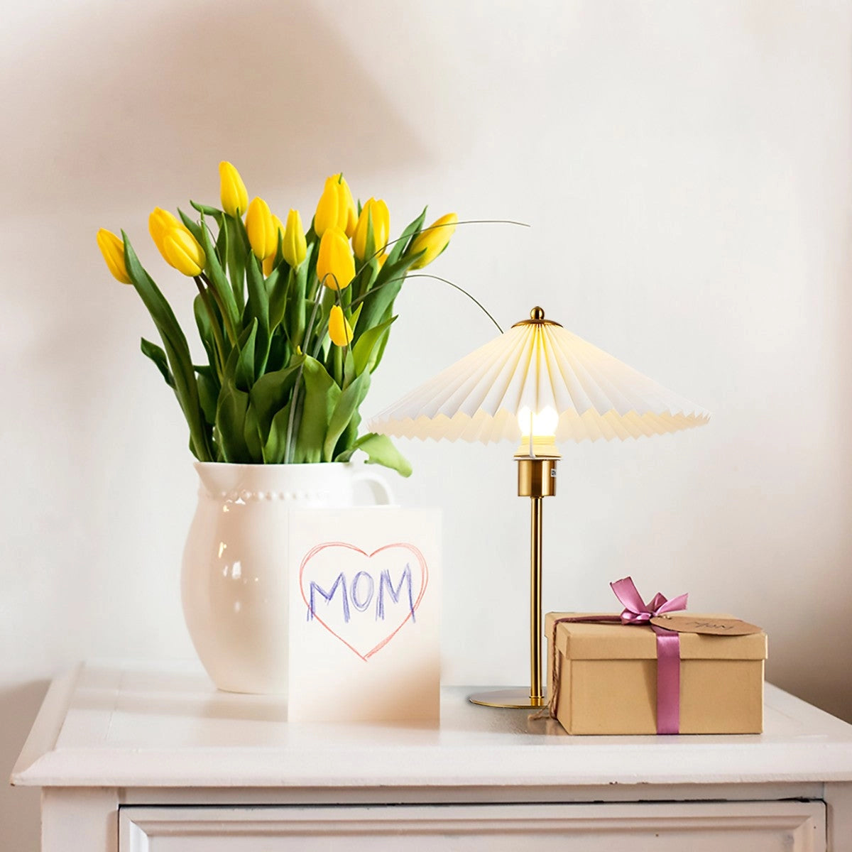 Modern gold pleated brass table lamp on a wooden table beside a yellow flowerpot with orange flowers and green leaves, enhancing the interior design of a bedroom or desk space.