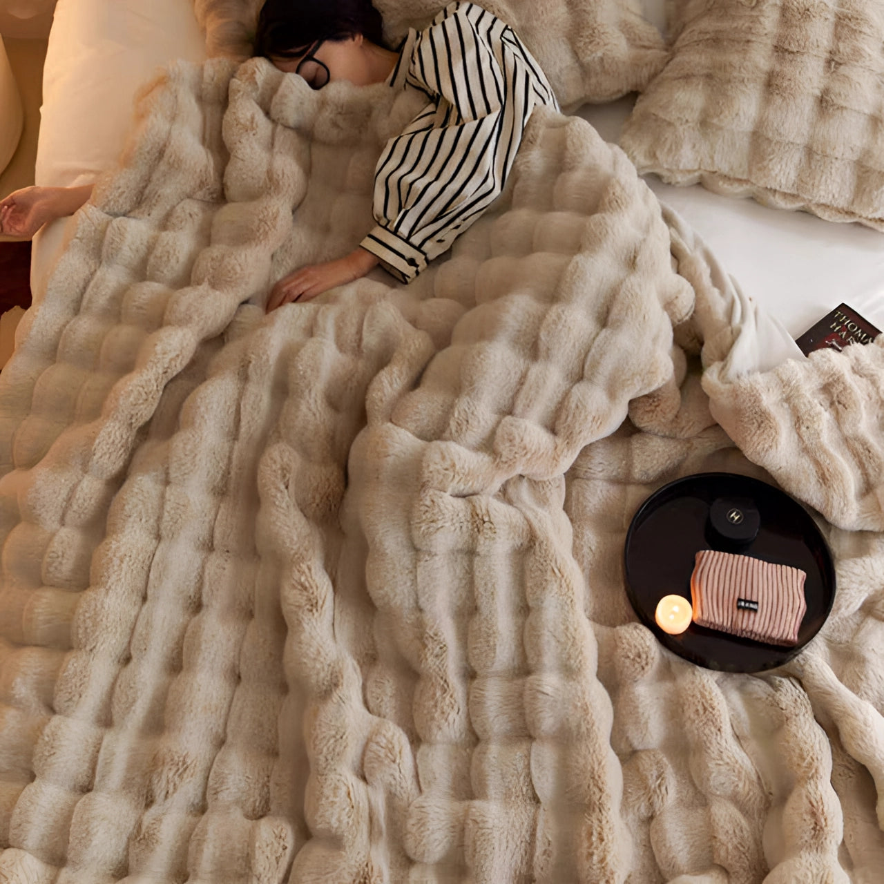 A plush pink chunky knit blanket draped over a couch, featuring a cozy and oversized design. The blanket exhibits a textured, woven pattern and is surrounded by wooden flooring and neutral-toned linens, enhancing the warm, comforting ambiance of the room. A camel-colored pillow rests alongside, complementing the overall aesthetic.