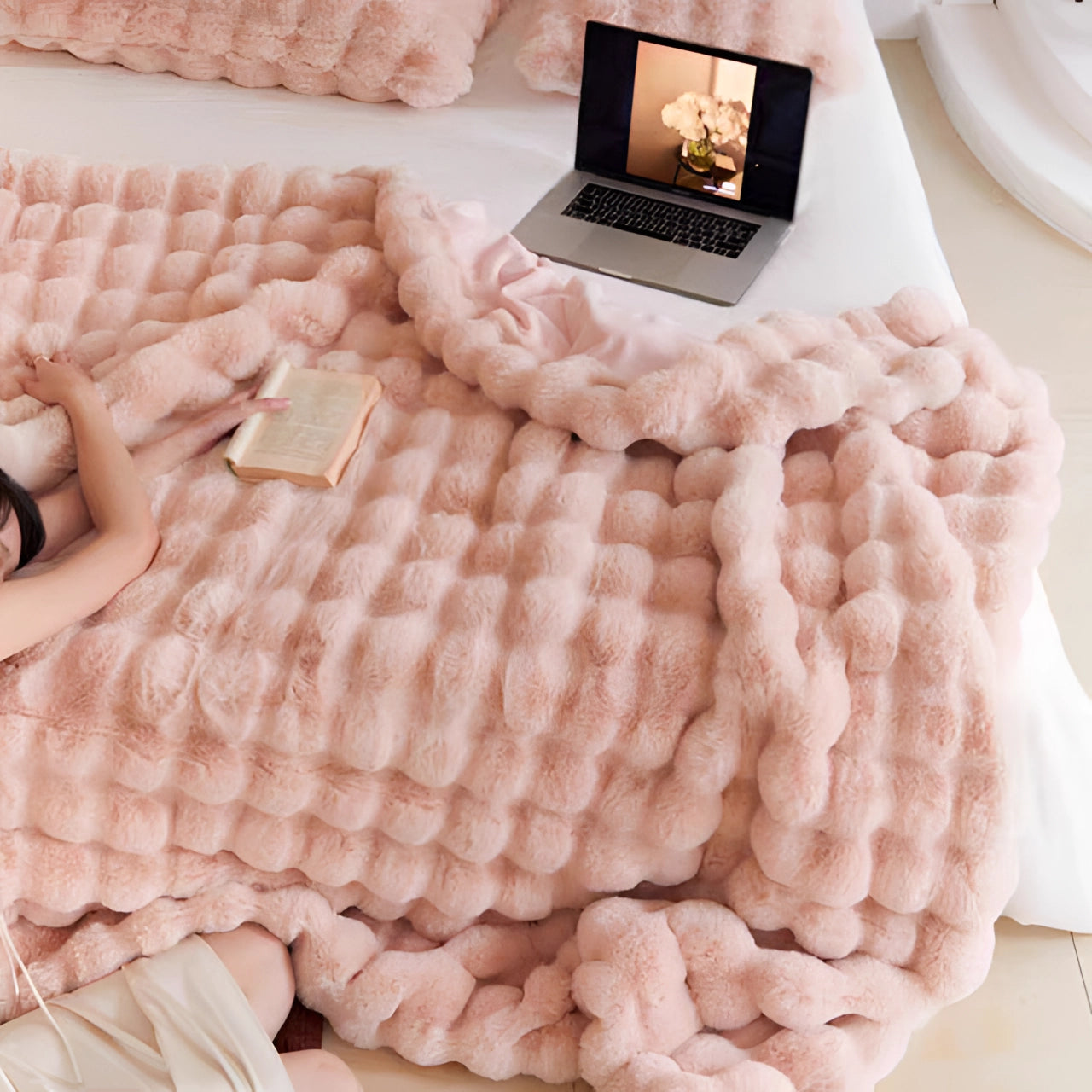 
An oversized plush pink chunky knit blanket draped over a cozy setting, featuring a wooden table with white tableware and a laptop nearby, with a pillow and linens on the floor.