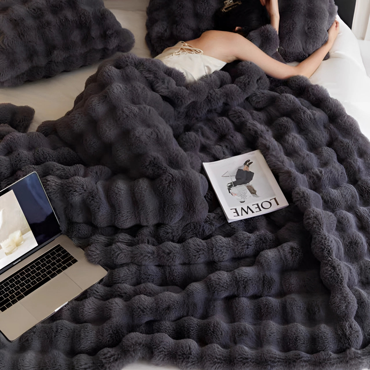 A dark gray chunky knit blanket draped over a couch with a laptop placed on top, creating a cozy and comfortable setting ideal for winter relaxation.