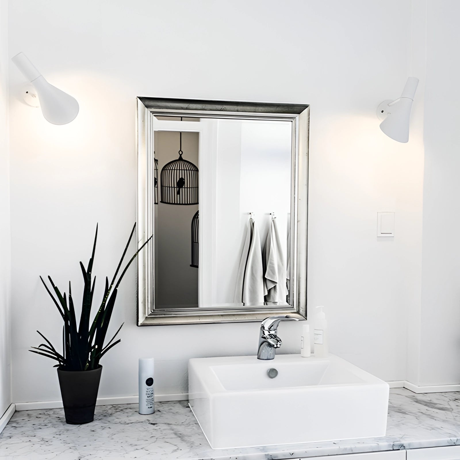 Rectangular silver mirror and wall light set in a modern bathroom setting featuring a white sink with a silver tap, a small green plant, and a combination of black and wood elements.