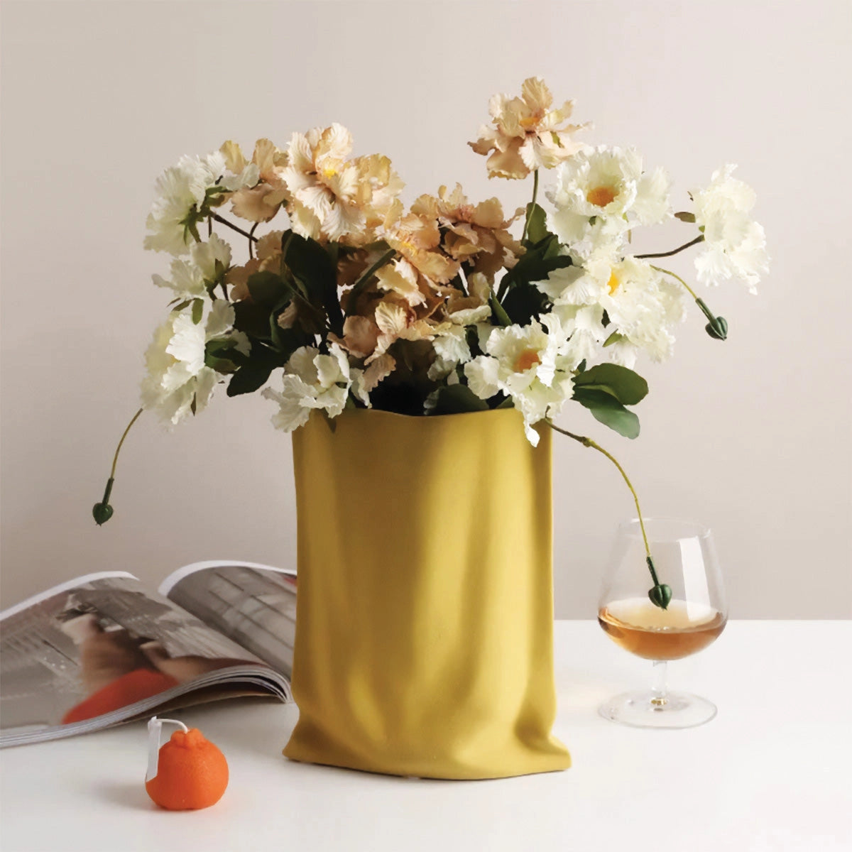 A modern reflective chrome vase in a high gloss yellow finish, showcasing a floral arrangement with various flowers and greenery, including branches and petals, elegantly displayed as a decorative piece.