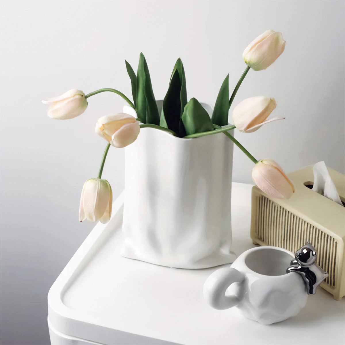 White reflective chrome vase with a modern high-gloss finish, styled with fresh flowers and petals, placed alongside dishware and drinkware on a table.