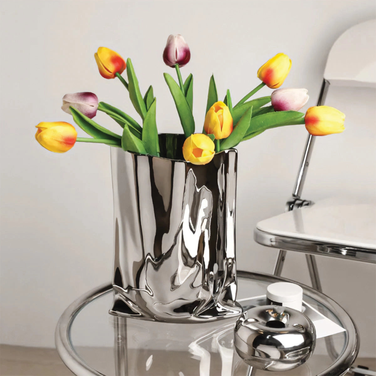 Reflective chrome vase with a modern high gloss finish, beautifully displaying orange flowers on a table, enhancing the interior design with a touch of elegance.