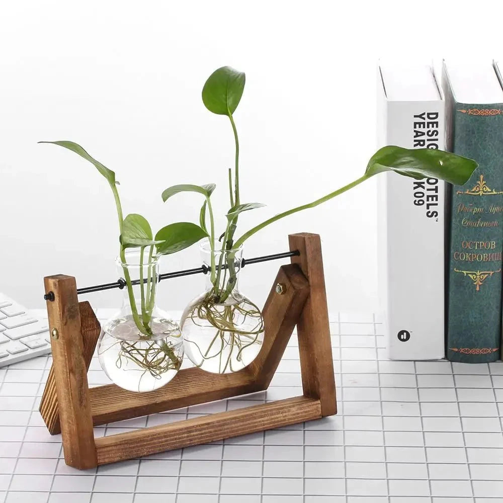 
A rustic wooden vase holder in a minimalist triangular design, featuring a glass container with a houseplant inside. The wooden stand appears to be crafted from hardwood and is placed on a surface, possibly outdoor furniture. The setting includes elements of nature, such as grass, complementing the earthy aesthetic of the vase holder.