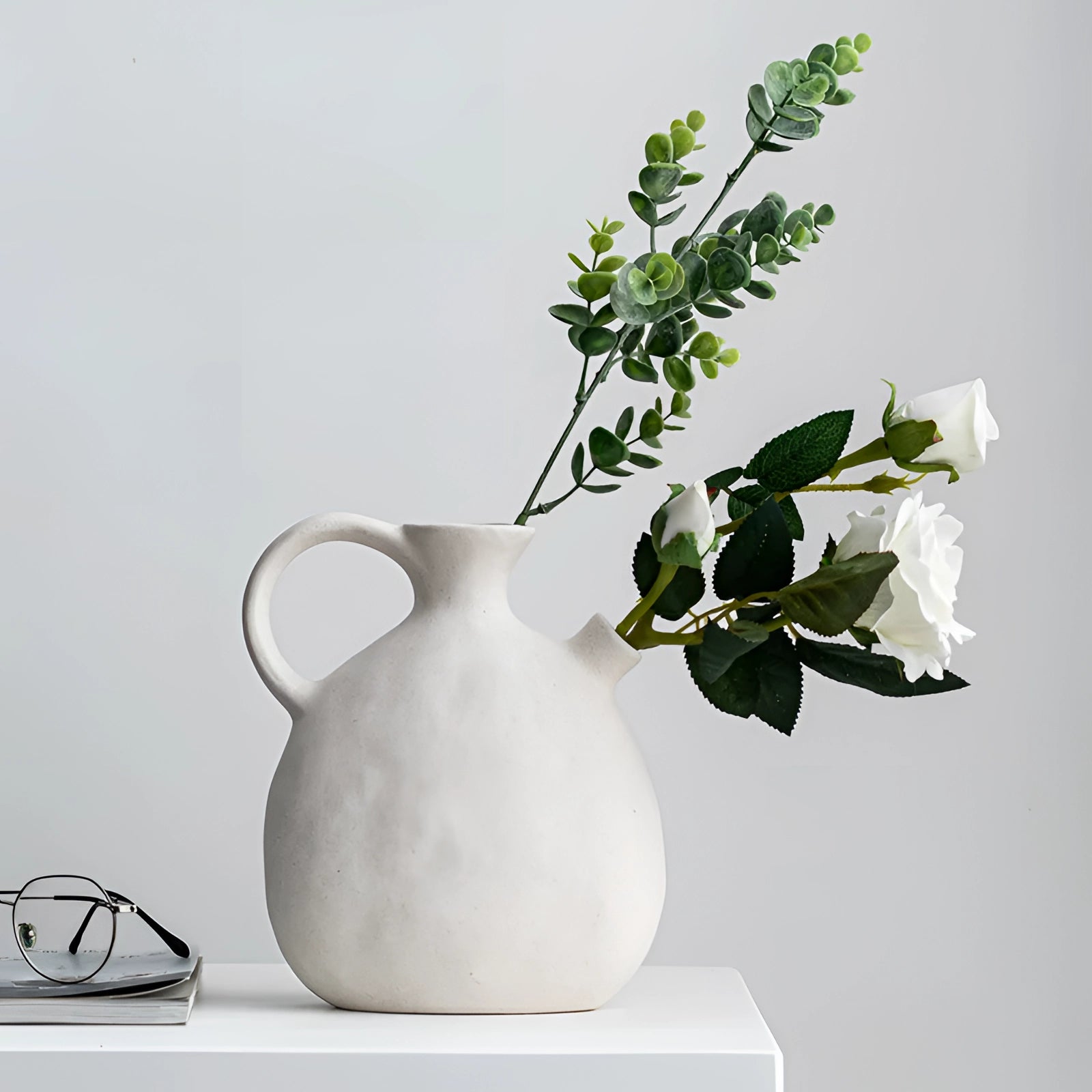 Scandinavian ceramic flower jug serving as a minimalist white vase with a simplistic design, holding a small arrangement of flowers, placed on a table.