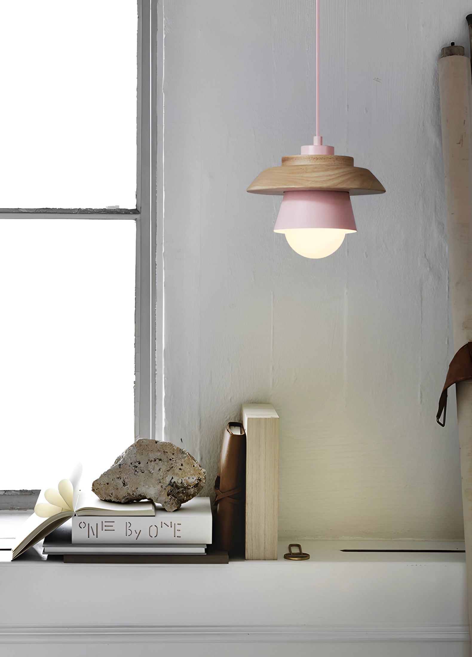 A modern Scandinavian-style wooden pendant light with a pink shade, hanging in a stylishly designed interior space with minimalistic decor elements, including windows, shelving, and linens.