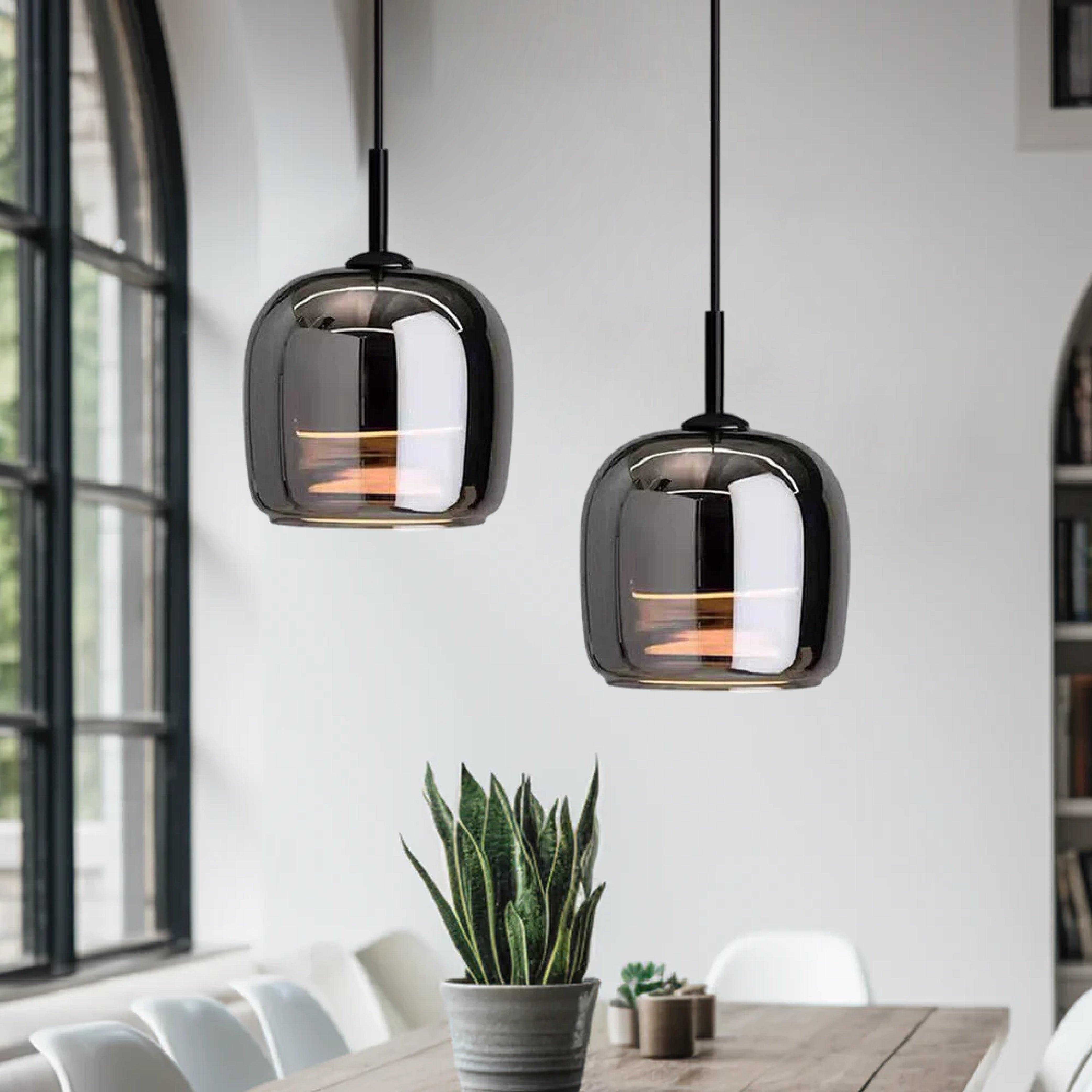 A smoked glass pendant light with a minimalist industrial design hanging above a kitchen island, featuring a sleek rectangular shape and subtle lighting, with wooden furniture and a plant in the background.