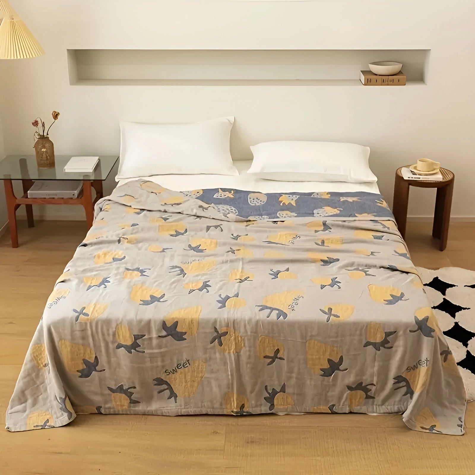 A cozy, grey strawberry-patterned blanket draped over a bed in a stylish bedroom, highlighting its comforting and decorative appeal against a backdrop of hardwood floors and modern furniture.