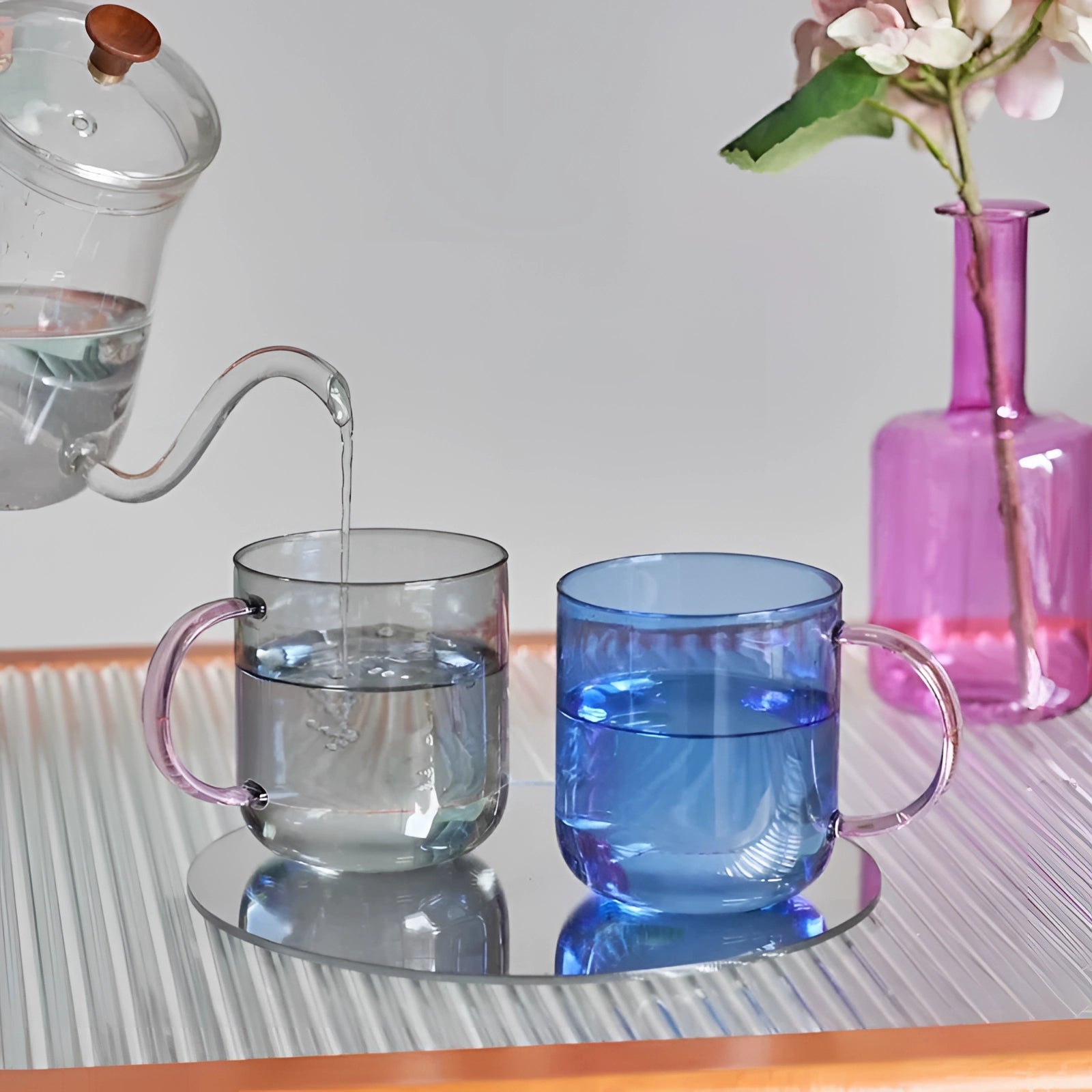 Stylish pink glass coffee and tea mugs filled with liquid, surrounded by a flower and a plant, showcasing elegant and unique glassware design.