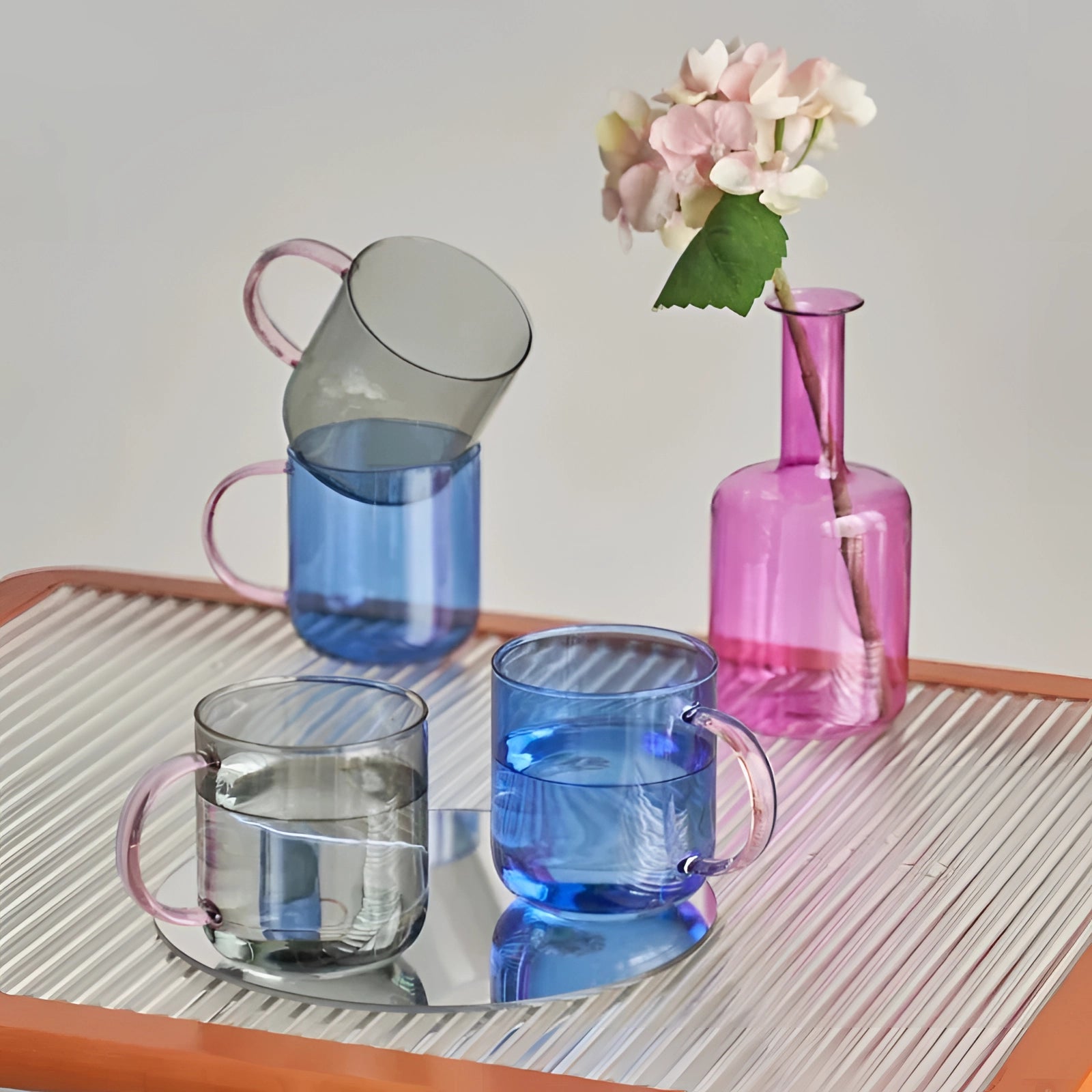 A set of stylish lightweight pink glass coffee and tea mugs featuring a unique design, displayed on a table alongside a decorative flower arrangement.