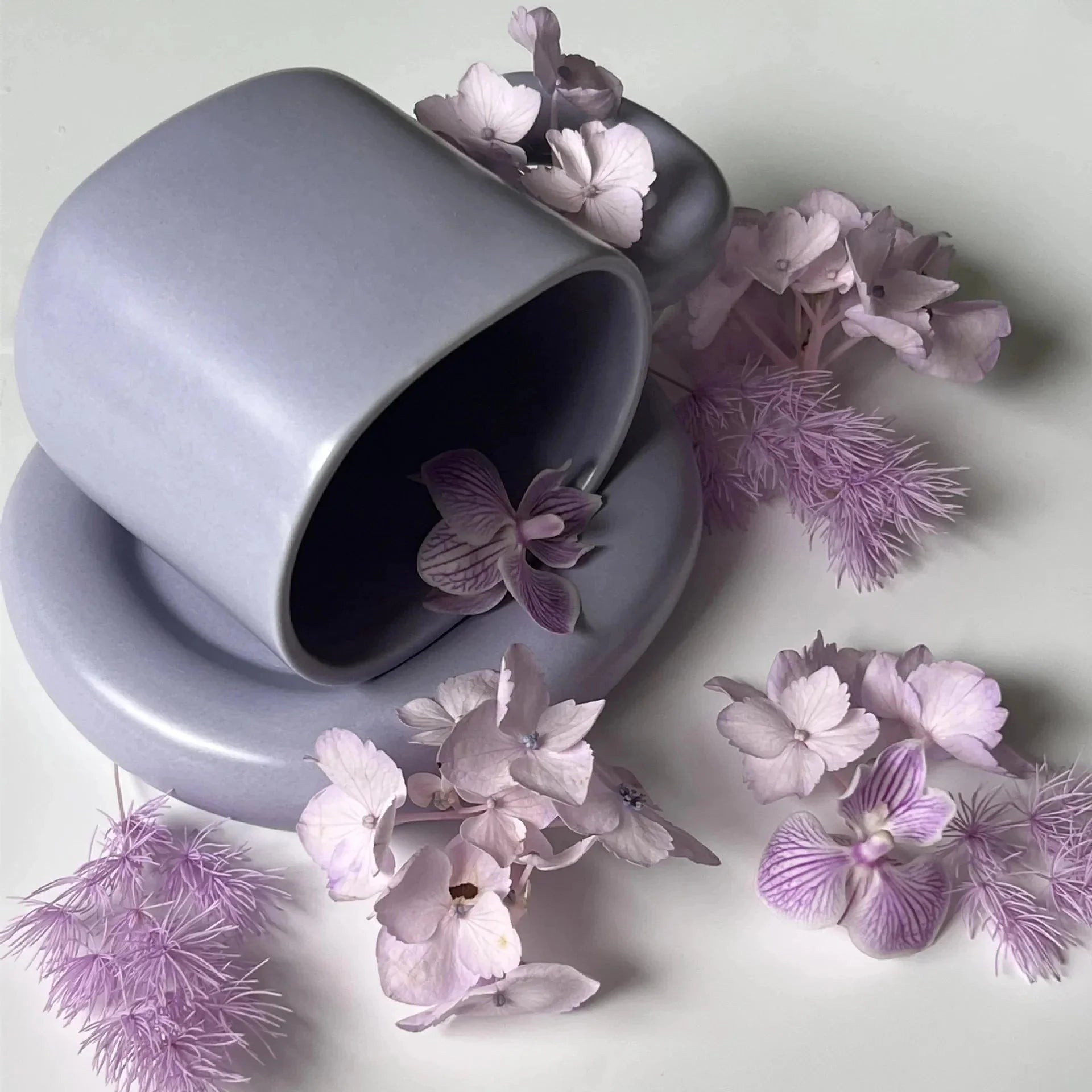 
A pale lilac ceramic mug with a unique handle and modern design, holding 280ml. The mug is surrounded by purple and pink flowers, contributing to a creative and artistic presentation.