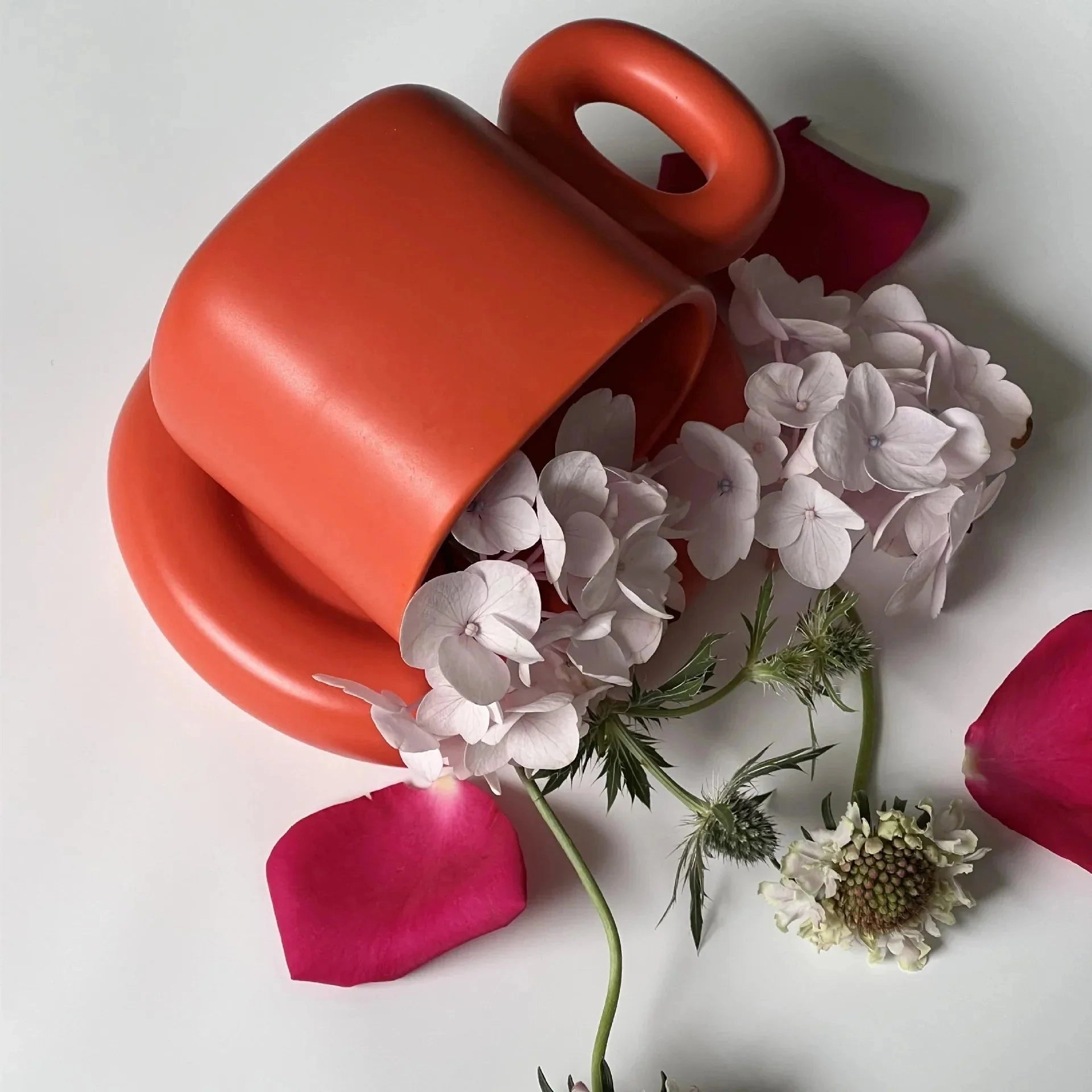 Turquoise ceramic mug with a unique handle, designed as a modern coffee cup, shown in an orange variant with a 280ml capacity. The mug is surrounded by colorful artificial flowers, including pink and magenta petals, arranged in a decorative bouquet.