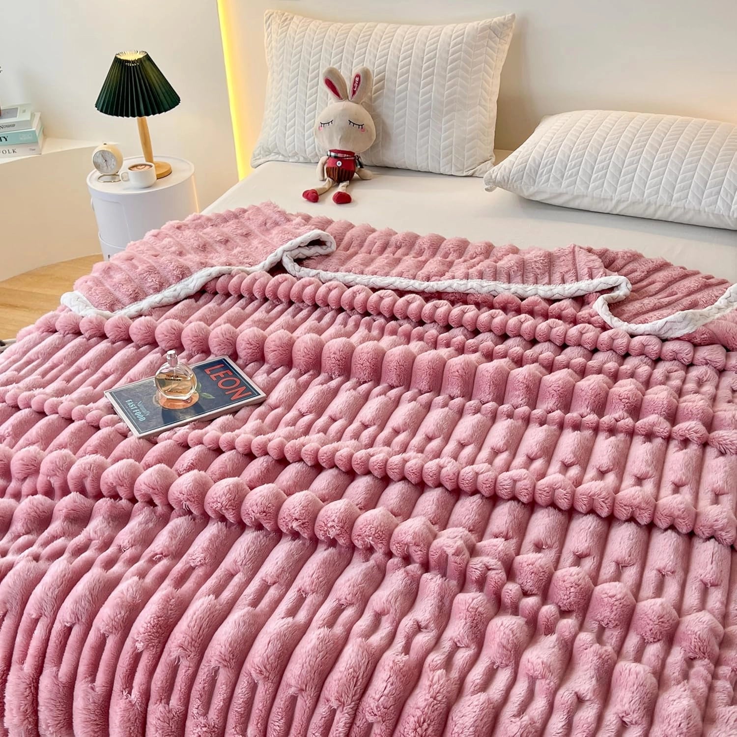 Pink chunky knit blanket draped over a bed, showcasing its luxurious braided texture. The blanket is set in a room with wooden flooring and a window in the background, highlighting its cozy and plush appearance.