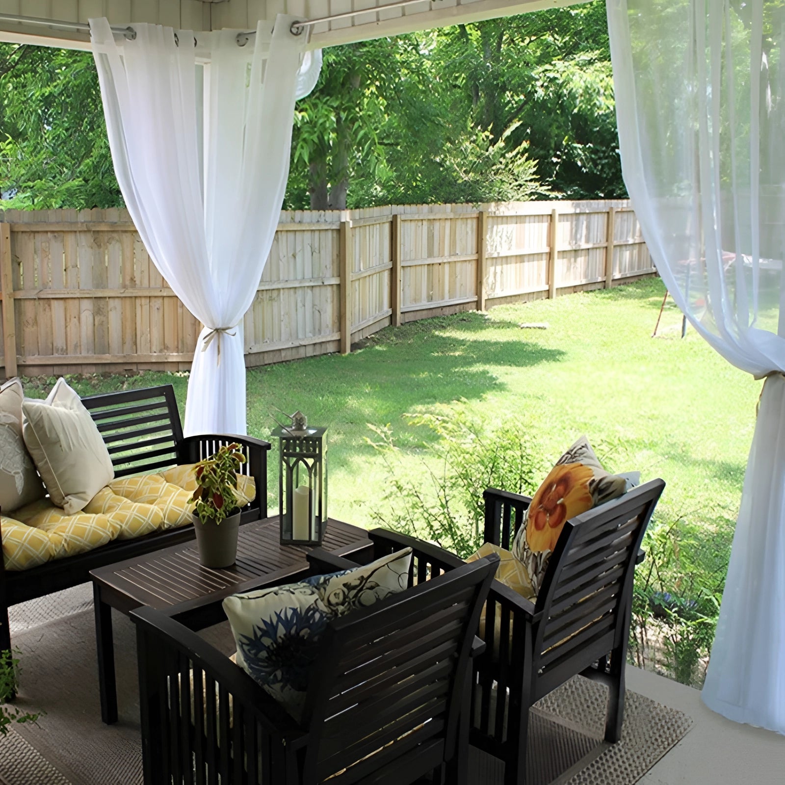 Waterproof sheer outdoor curtains in white, elegantly draped around an outdoor seating area with a table and chairs, surrounded by greenery and a building in the background.