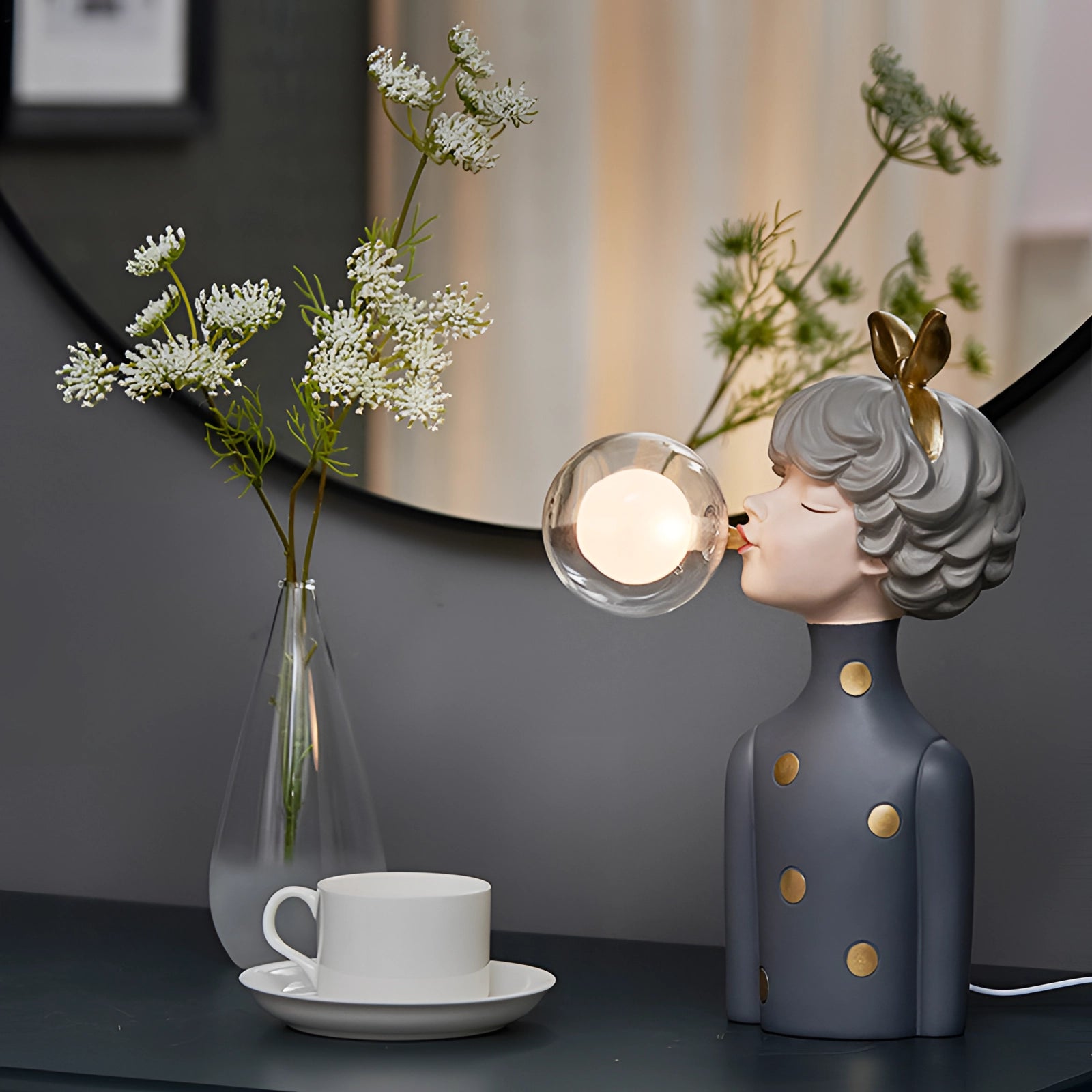 A whimsical ceramic bubble statue featuring gold bunny ears, displayed alongside a branch-like plant on a table in a well-lit interior setting.